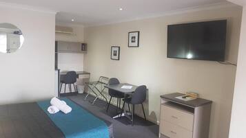 Quarto de hotel com cama, escrivaninha, cadeira, televisão e uma pequena cozinha.