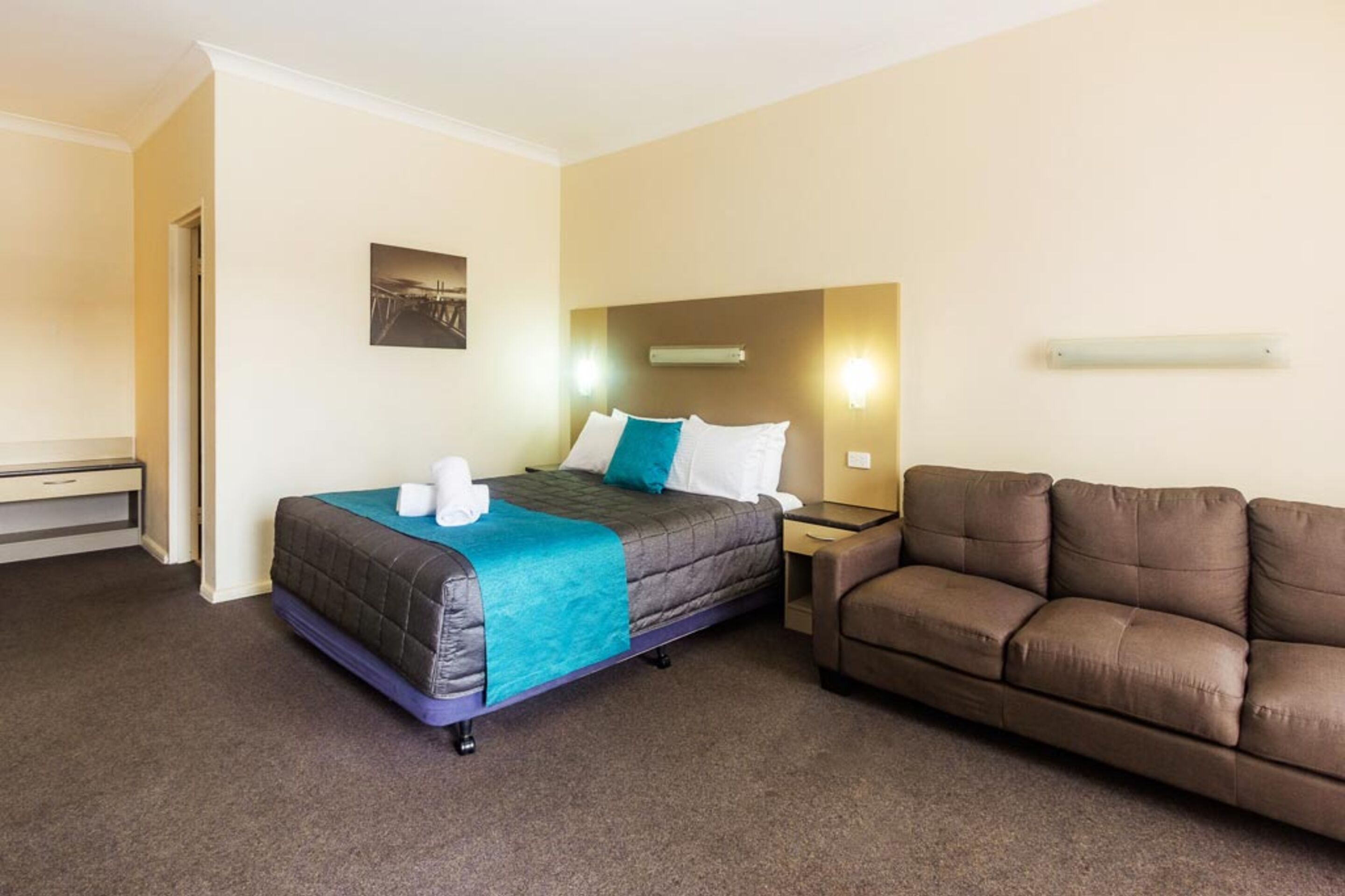 Quarto de hotel com cama, sofá marrom, mesa de cabeceira e luminária fixada na parede.