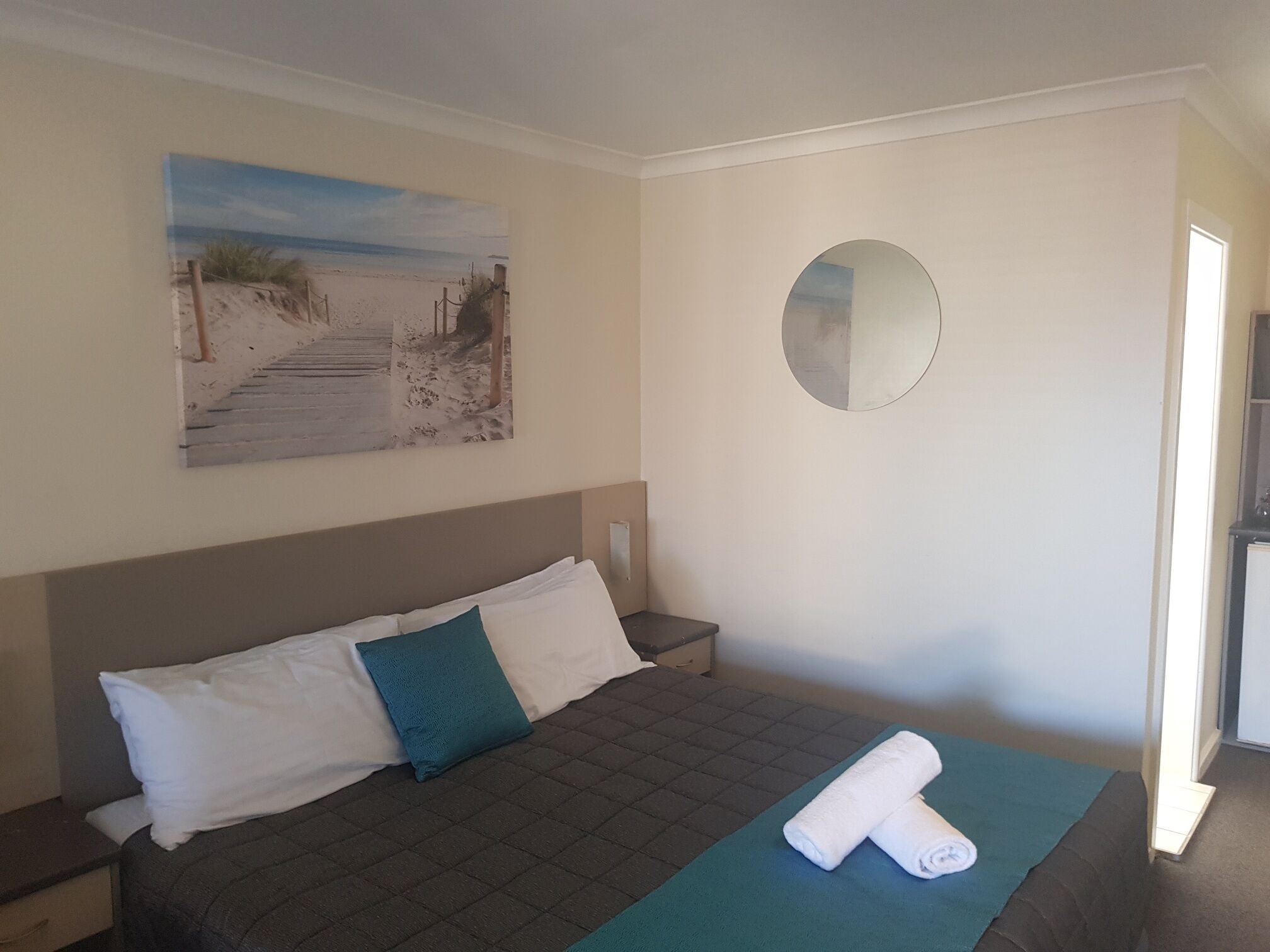 Una camera d'albergo con un letto, un dipinto di una spiaggia, uno specchio tondo e un letto rifatto con un asciugamano.