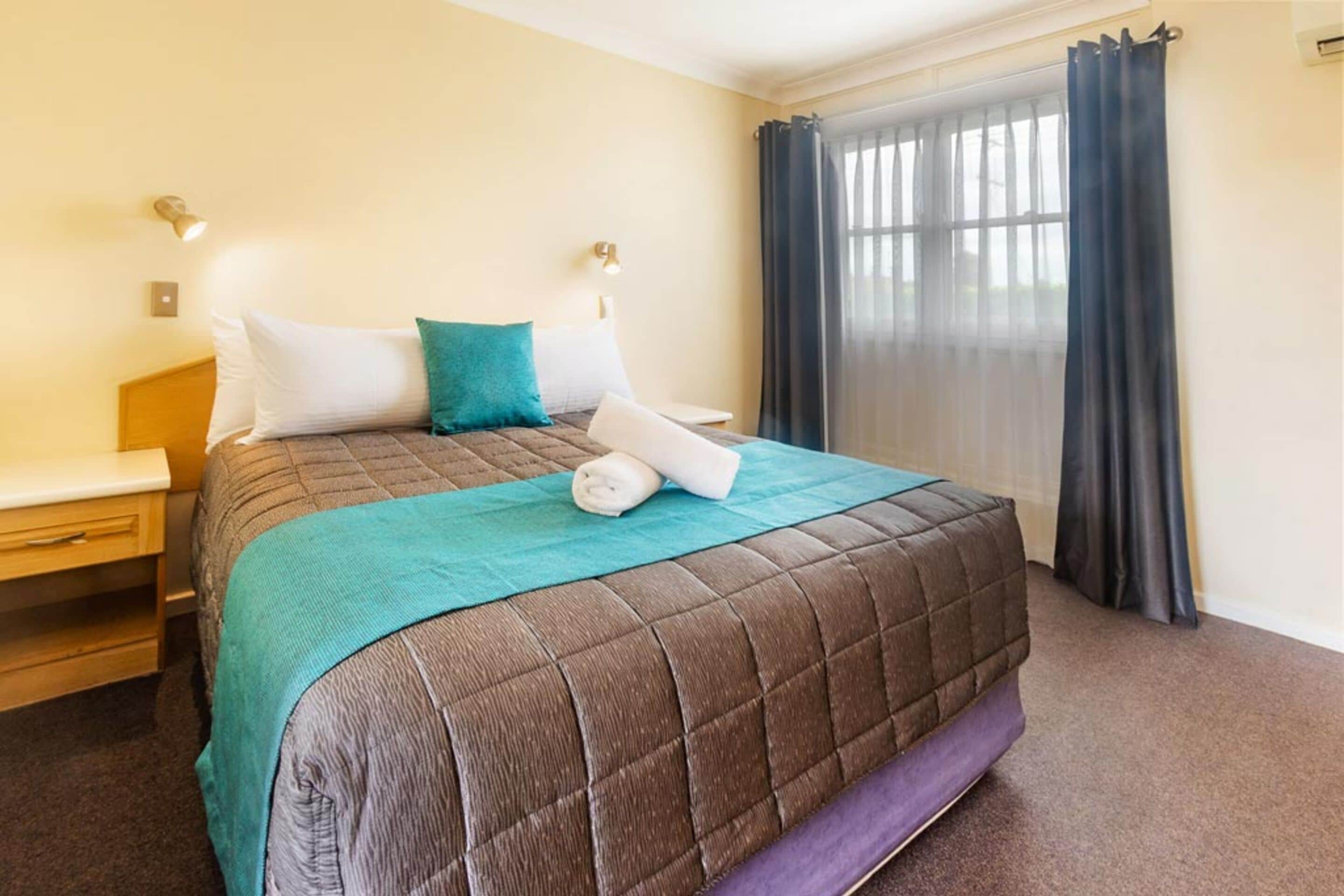 A neatly made bed with a brown quilt, a teal throw blanket, and a rolled towel. There are two bedside tables with lamps, a window with dark curtains, and a wooden nightstand.