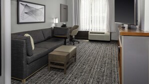 Et moderne hotelværelse med en grå sofa, en lille træpuf, et skrivebord med stol, et fladskærms-tv og et stort vindue med lette gardiner.