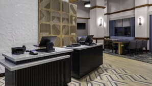 Et hotellets receptionsområde med to receptionister, en stor vægpanel med geometriske mønstre, et siddeområde med et bord og stole, samt et stort vindue med gardiner.
