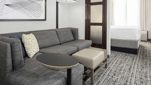 Et moderne hotelværelse med en grå sofa, et lille rundt bord, en seng med hvide sengetæpper og et stort vindue med lodrette persienner.