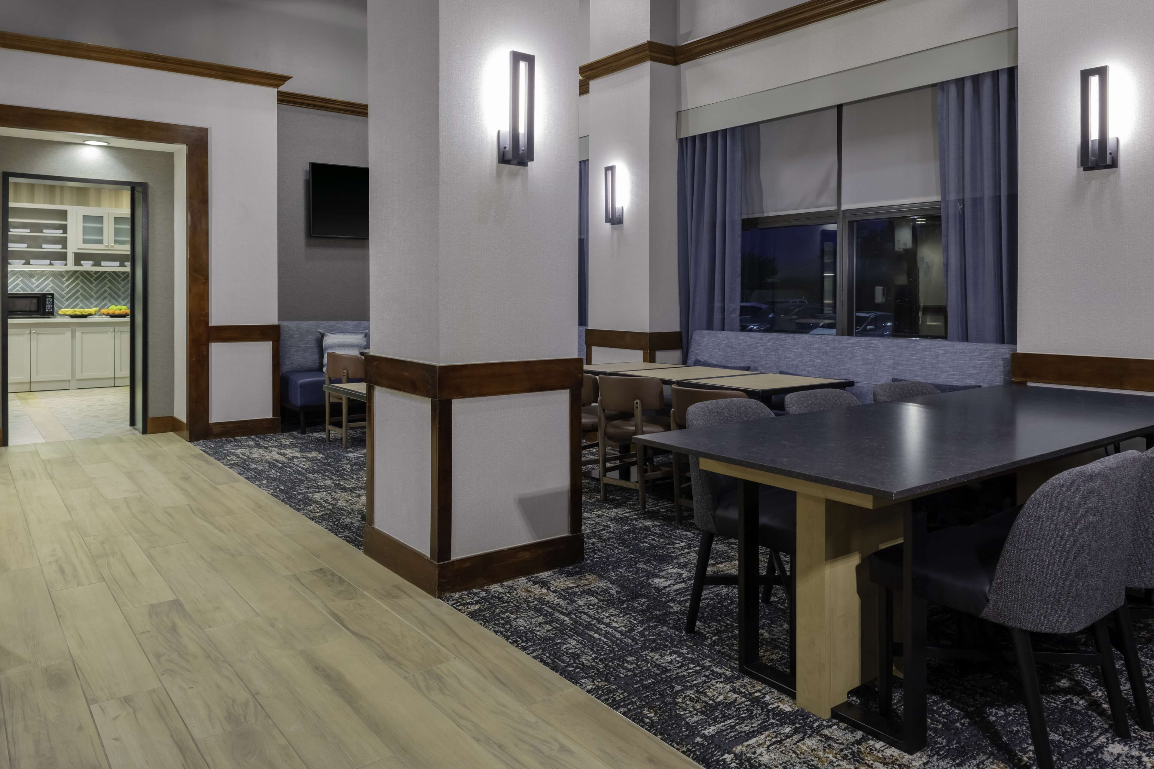 A hotel room with a dining area, a kitchen in the background, and a large window with curtains.