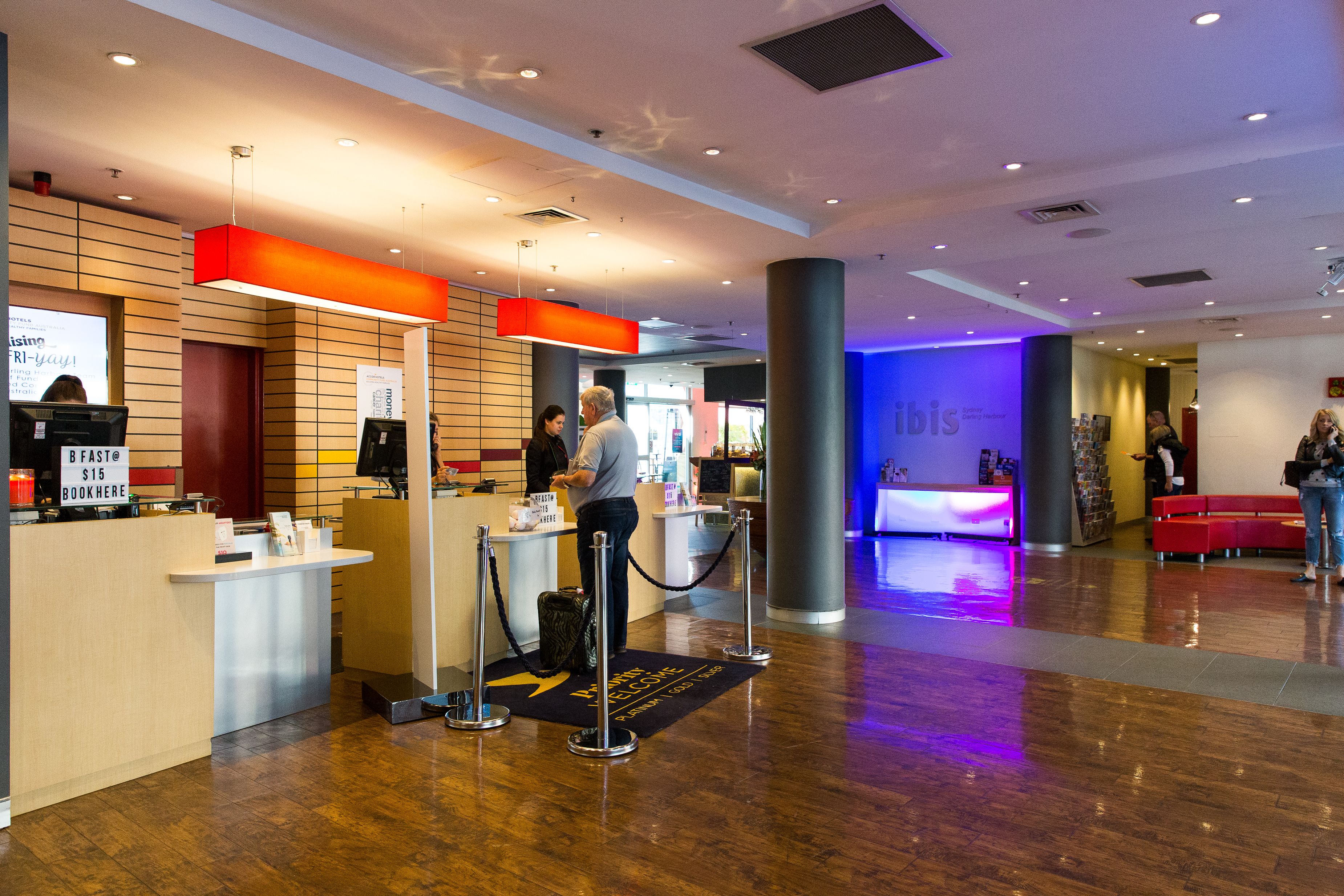 Zona de recepción de hotel con diseño moderno, que incluye un mostrador, una pantalla digital y un área de estar con sofás rojos.