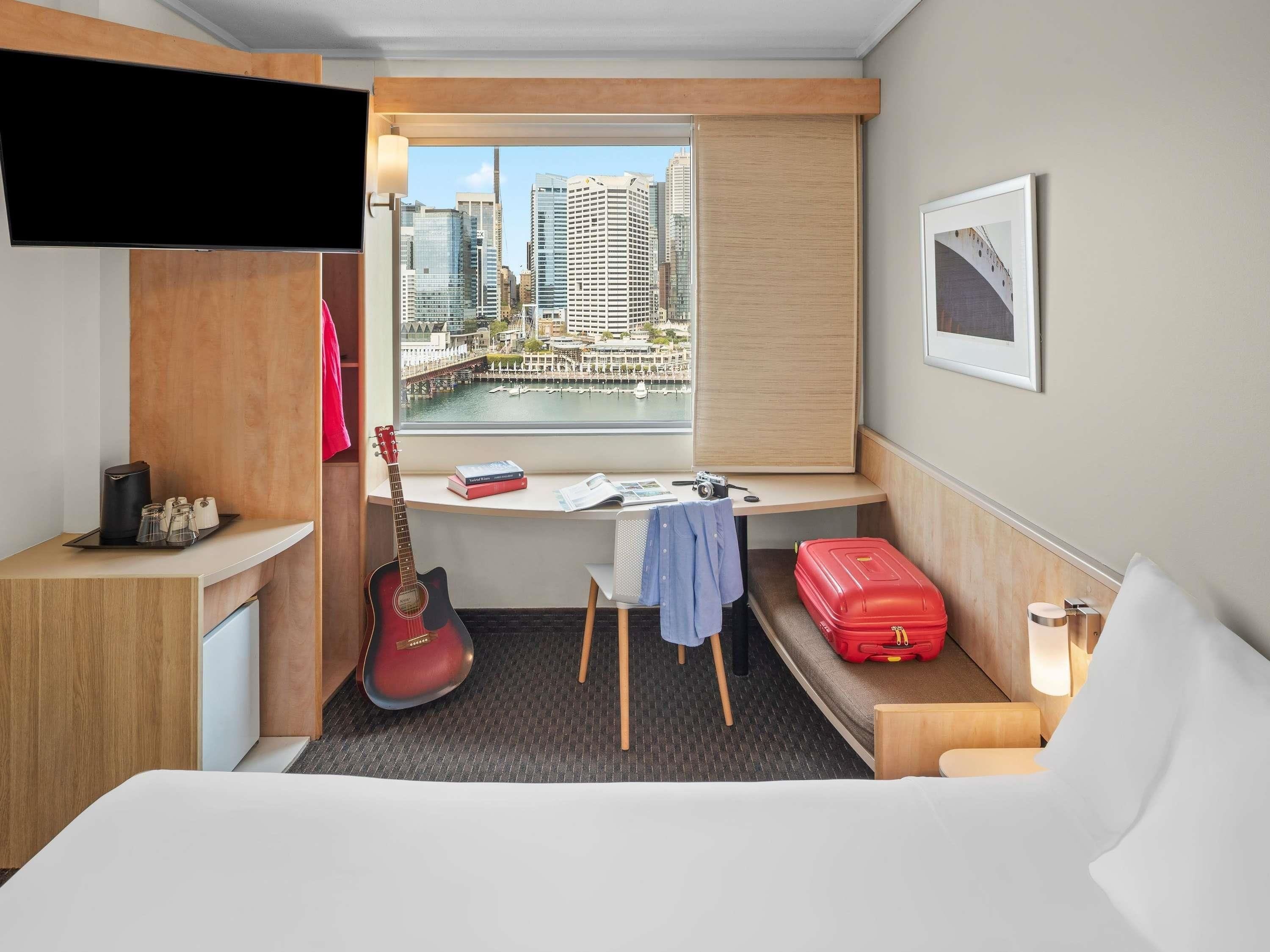 A hotel room with a desk, a guitar, a red suitcase, and a view of a cityscape.