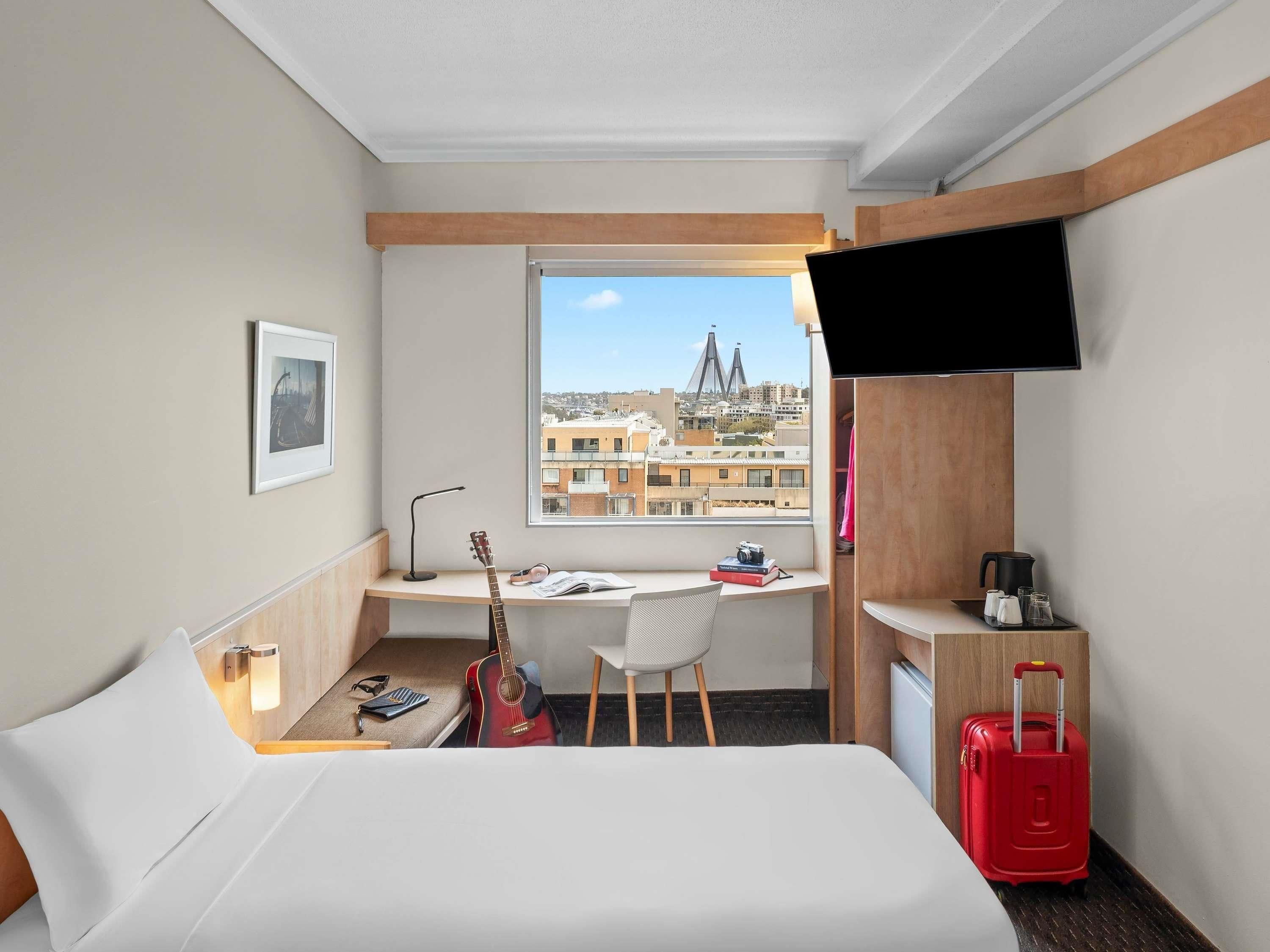 A hotel room with a bed, a desk with a guitar, a chair, a TV, and a window with a city view.