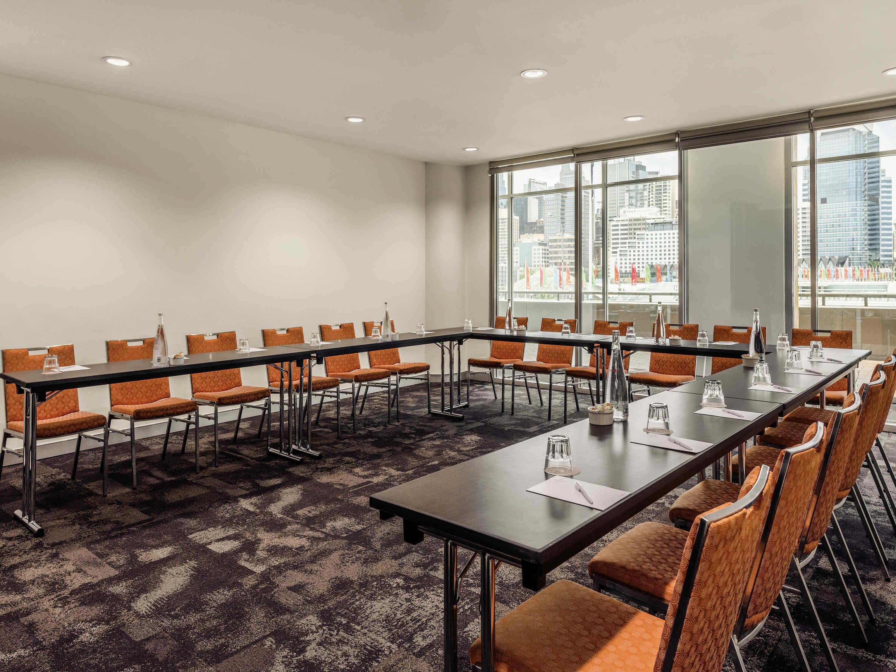 Une salle de conférence dotée de plusieurs tables et chaises, de grandes fenêtres offrant une vue sur la ville et d’un sol recouvert de moquette.