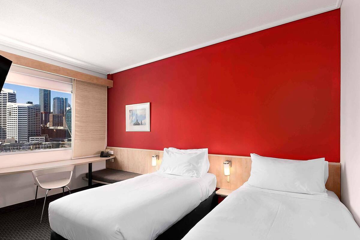 A hotel room with two beds, a desk, a chair, a city view, and a red accent wall.