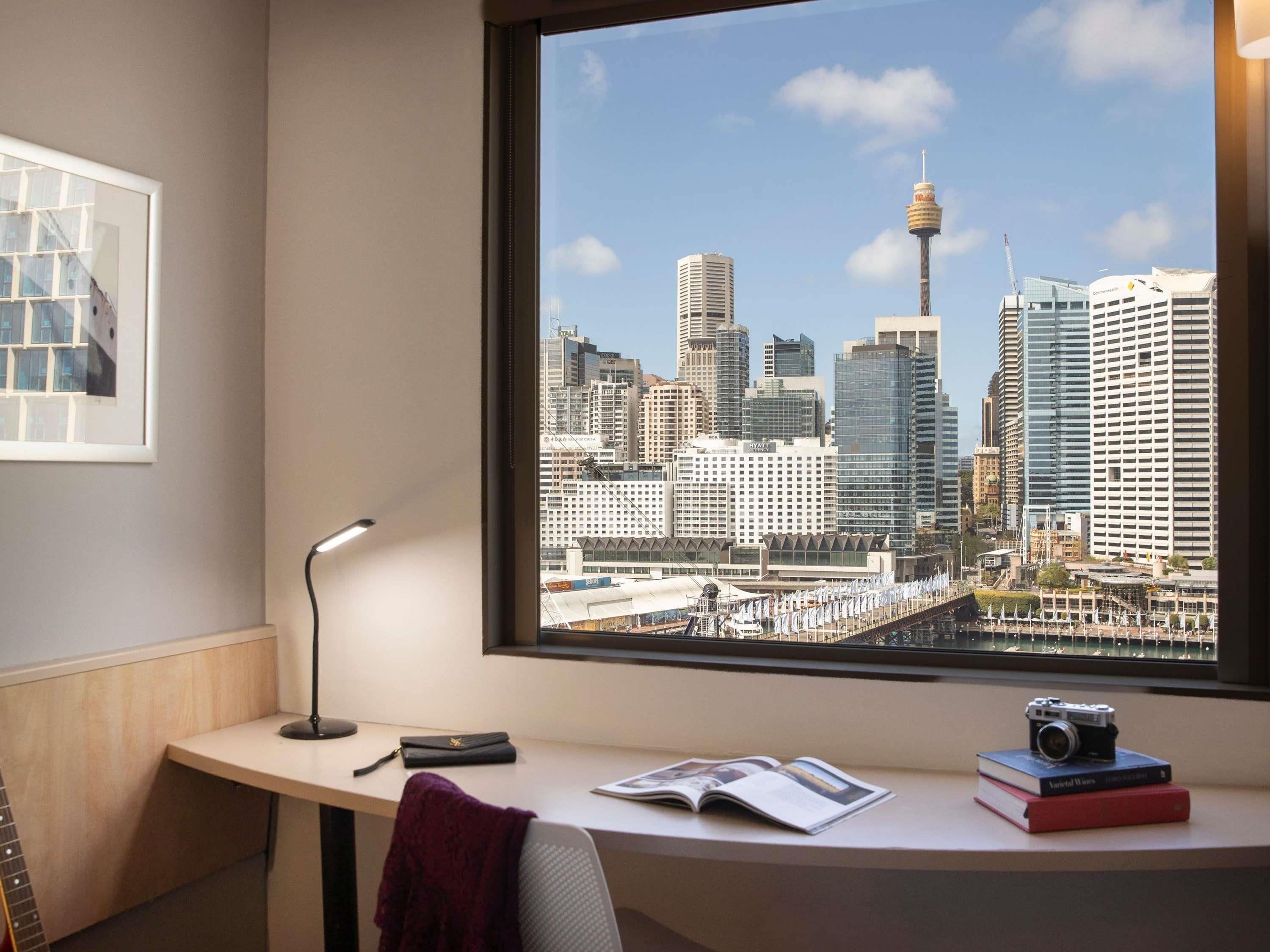 Un escritorio con una cámara, libros y una lámpara, con vistas a un paisaje urbano con edificios modernos y una torre.