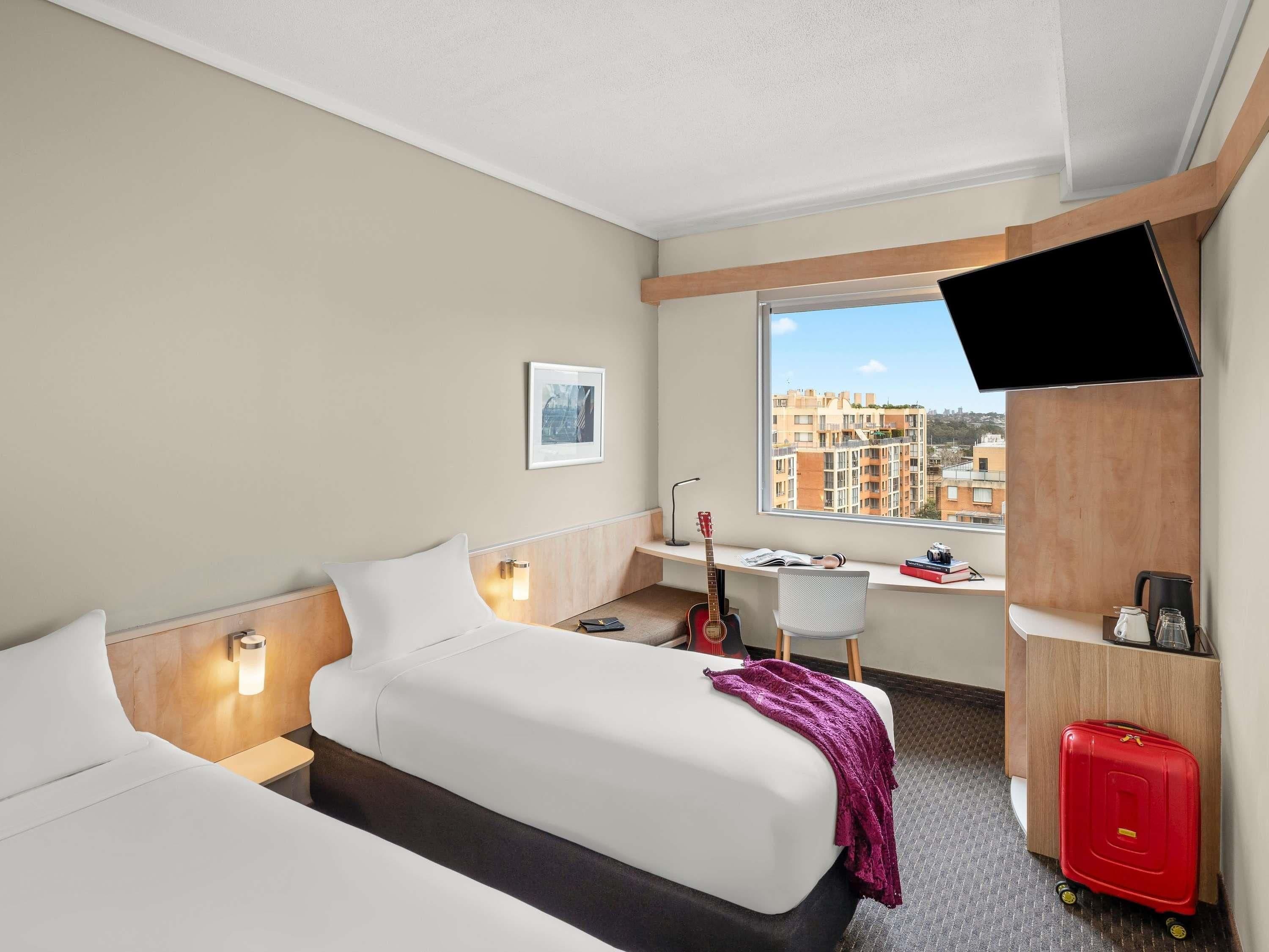 Une chambre d’hôtel avec un lit, un bureau, une chaise, une guitare, une valise et un téléviseur.