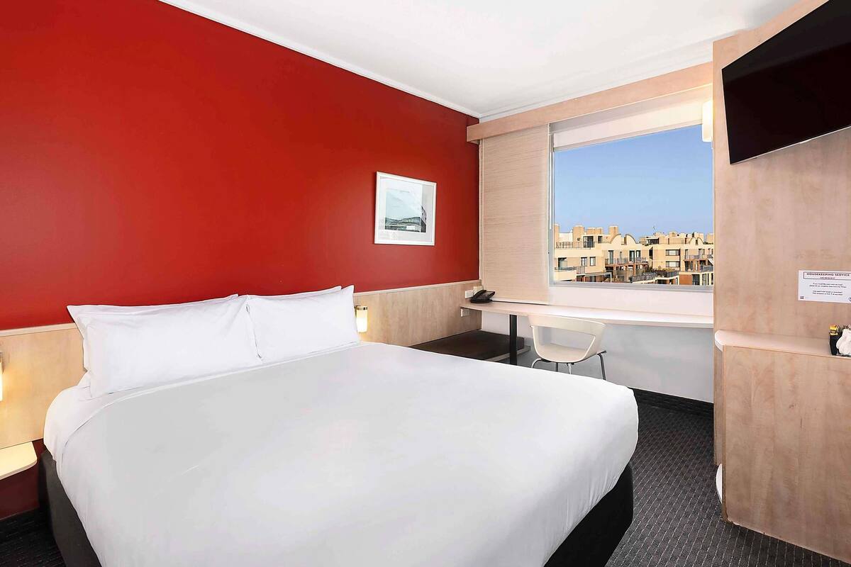 A hotel room with a large bed, a desk, and a chair. There is a window with a view of buildings outside.