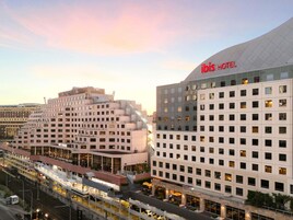 A modern hotel with a prominent 'ibis HOTEL' sign, multiple floors, and a central courtyard with outdoor seating.