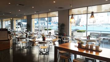 A spacious restaurant with high ceilings and large windows offering views of a cityscape and waterfront. Tables and chairs are arranged for dining.
