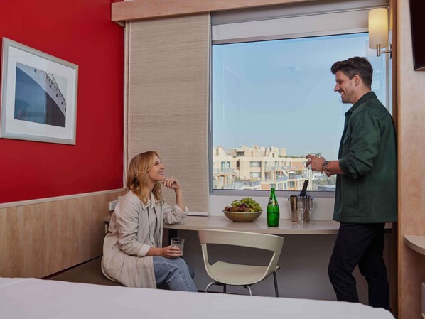 Una habitación con pared roja, ventana con vista a la ciudad, una mesa con un frutero, una botella y un vaso.