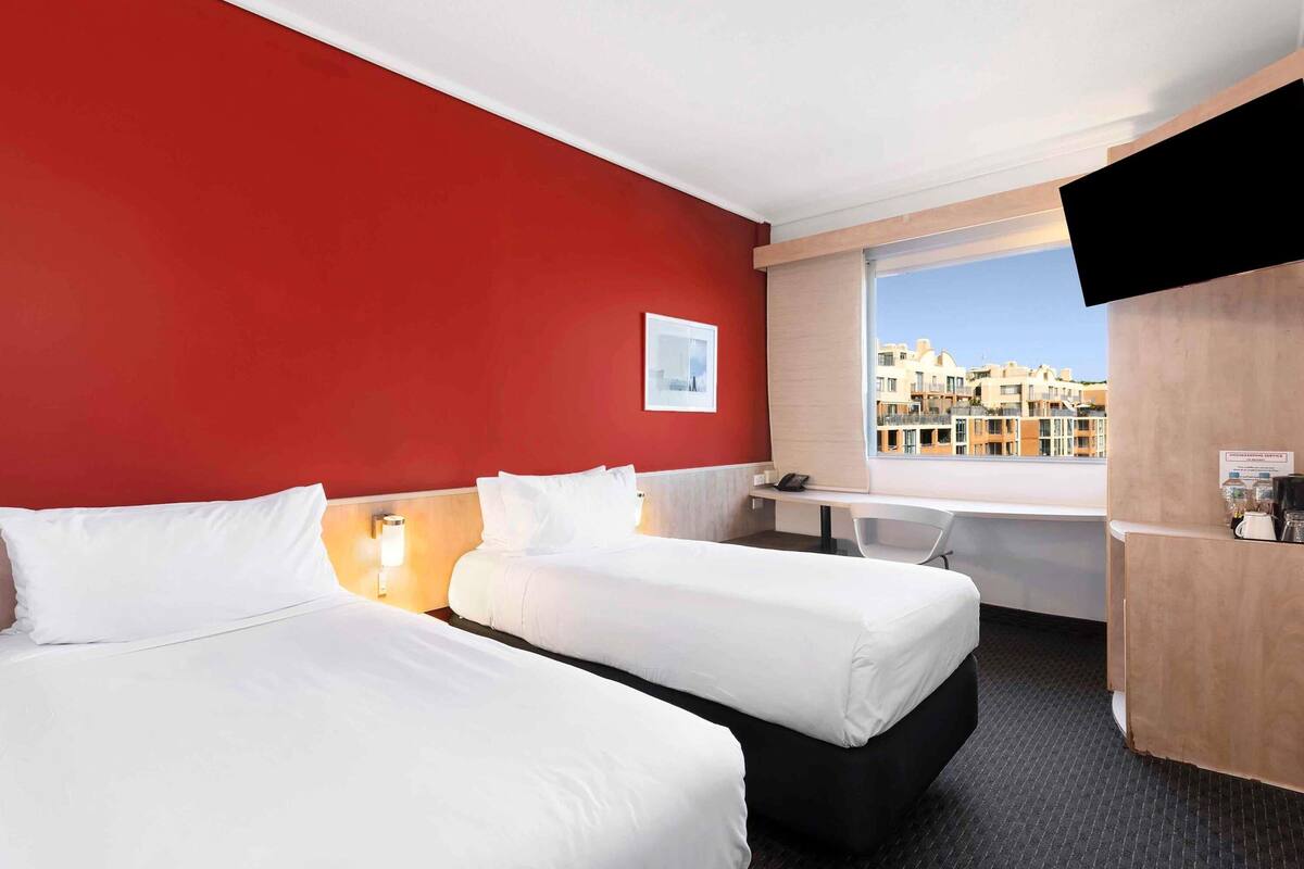 A hotel room with two beds, a desk, a chair, a TV, and a window with a city view.