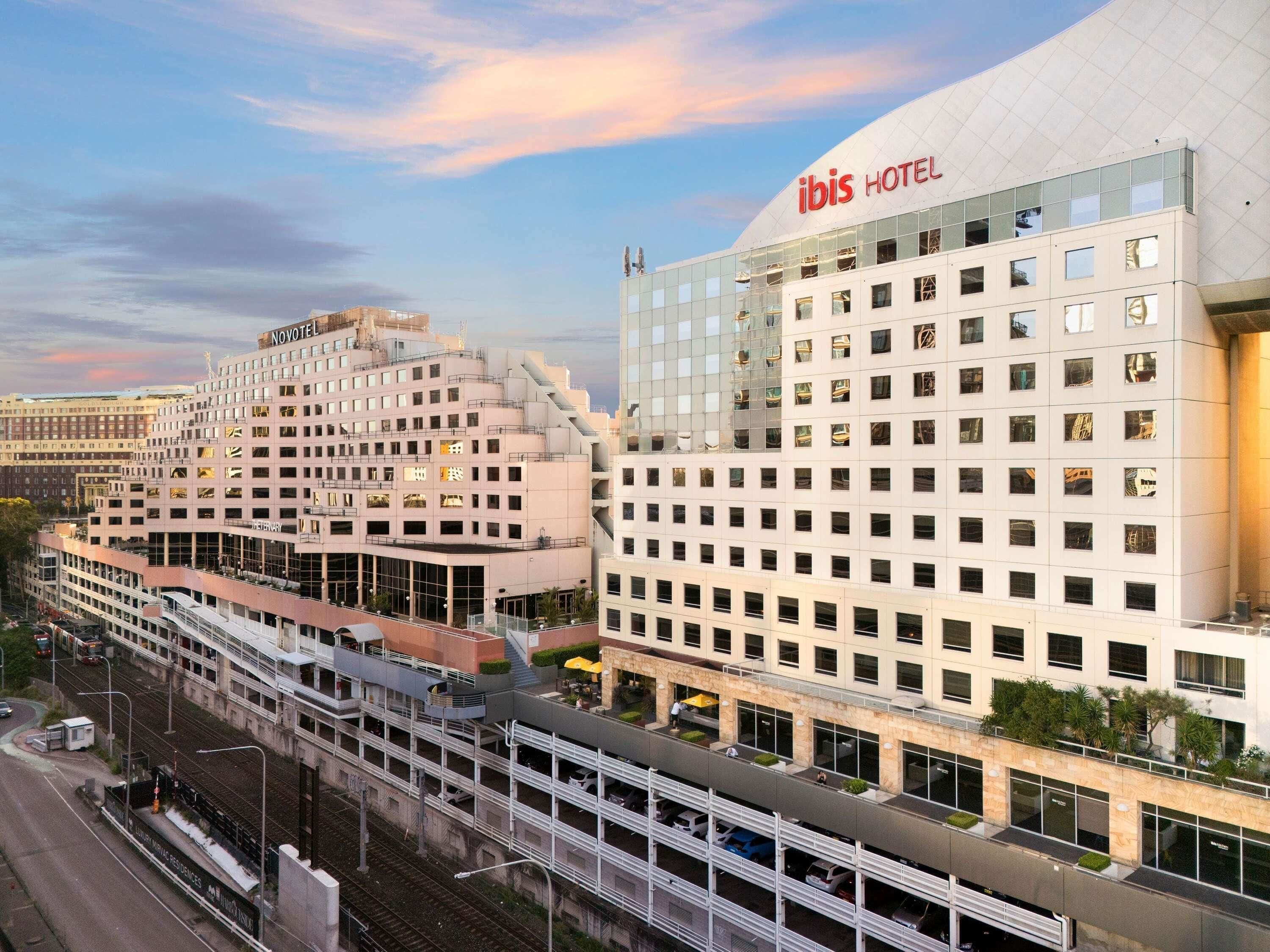 Un hôtel moderne, reconnaissable à son grand panneau « IBIS HOTEL », une structure à plusieurs étages dotée d’un toit plat et un espace de stationnement agrémenté d’une allée couverte.