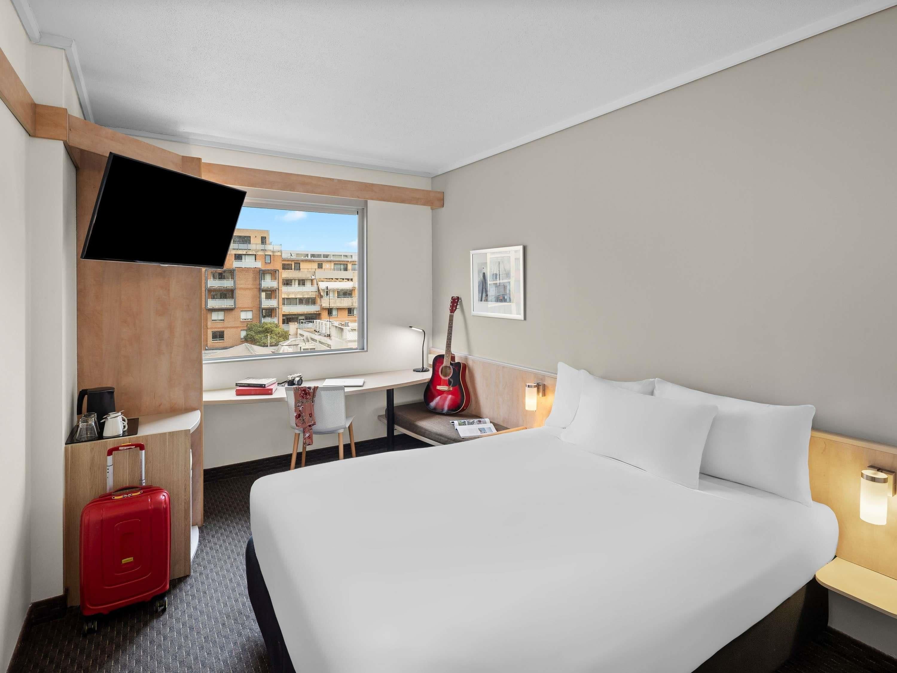 A hotel room with a bed, a desk, a guitar, a TV, and a window with a city view.