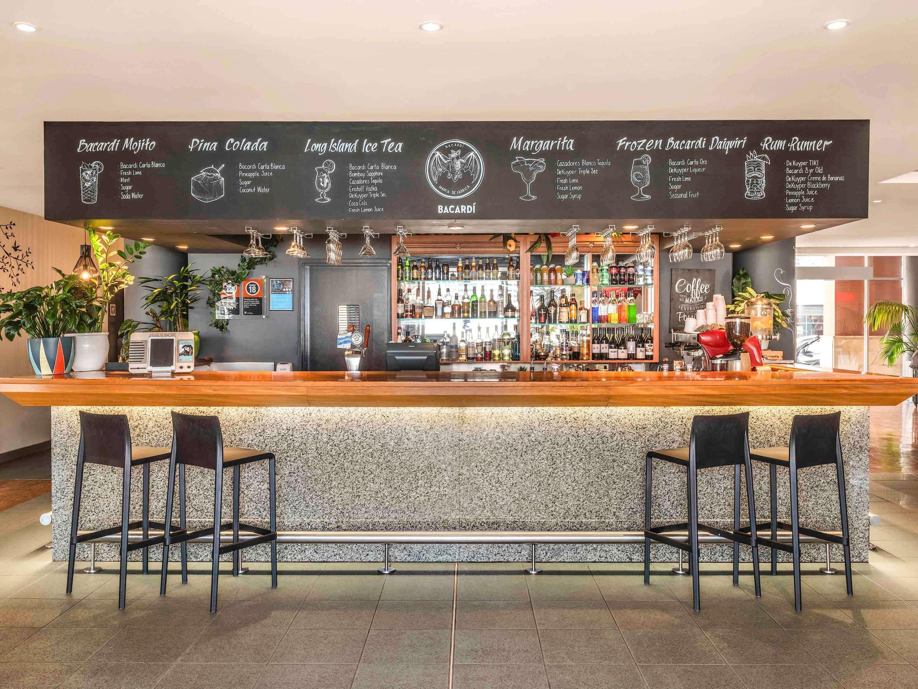 A bar area with a menu board, bar stools, a cash register, and a variety of bottles and glasses.