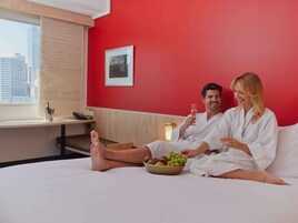 Habitación de hotel con una pared roja, una cama con ropa de cama blanca, un frutero y una ventana con vistas a la ciudad.