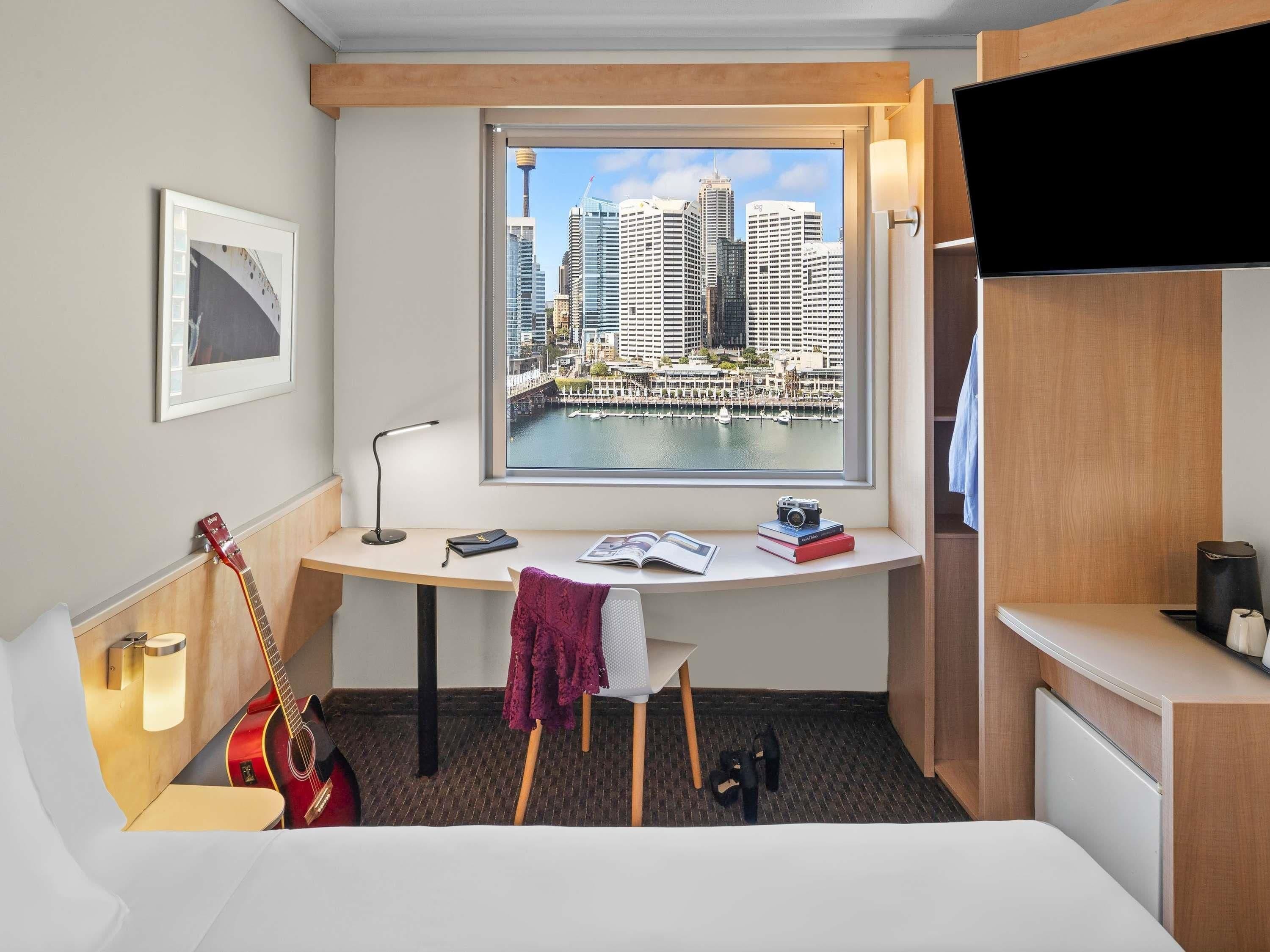 Een hotelkamer met een bureau, een gitaar, een stoel, een bed en uitzicht op een stadsbeeld door een raam.
