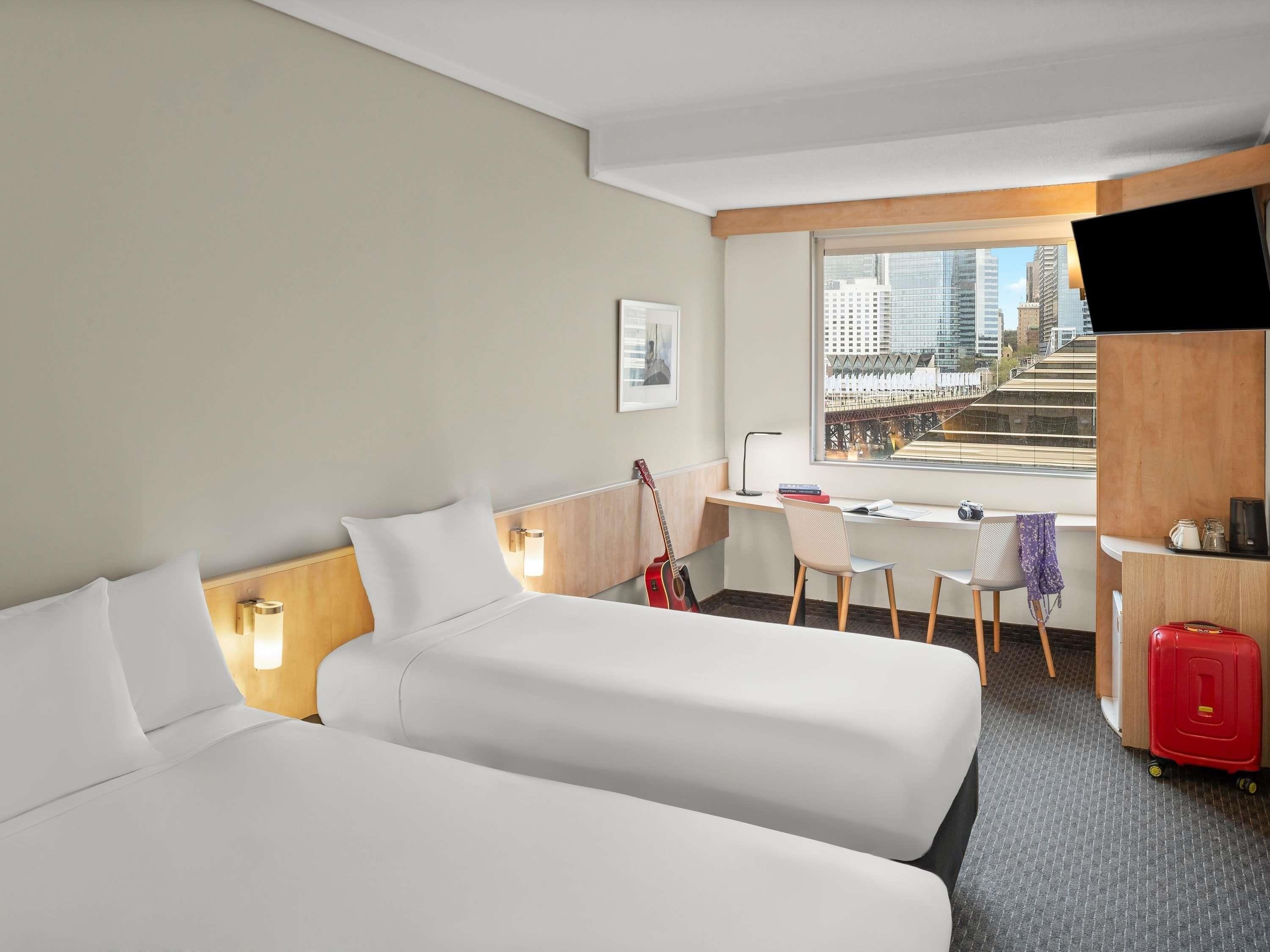 A hotel room with two beds, a desk, a chair, a guitar, a suitcase, and a city view.
