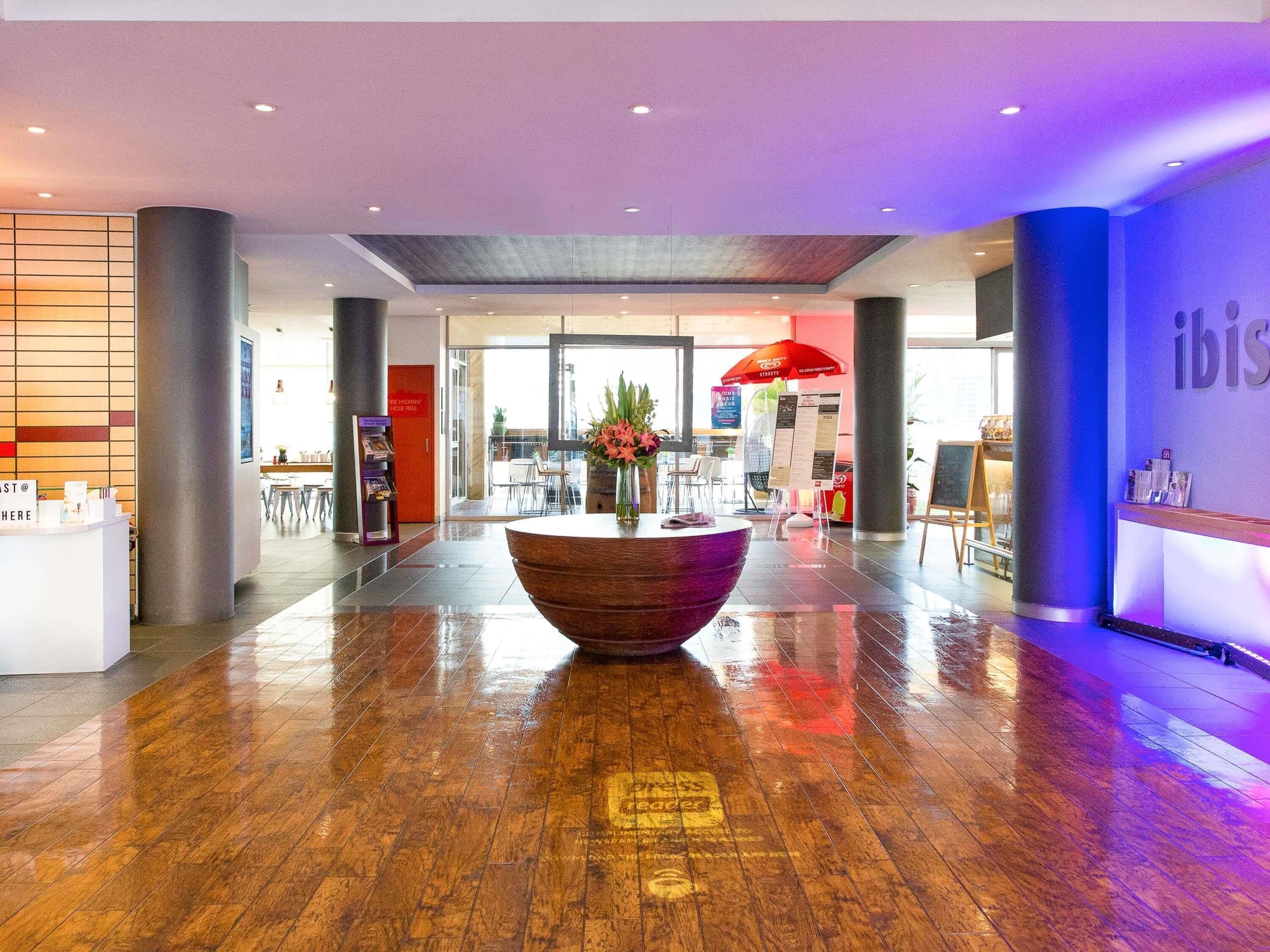 A modern hotel lobby with a wooden reception desk, a large flower vase, and a seating area with tables and chairs.