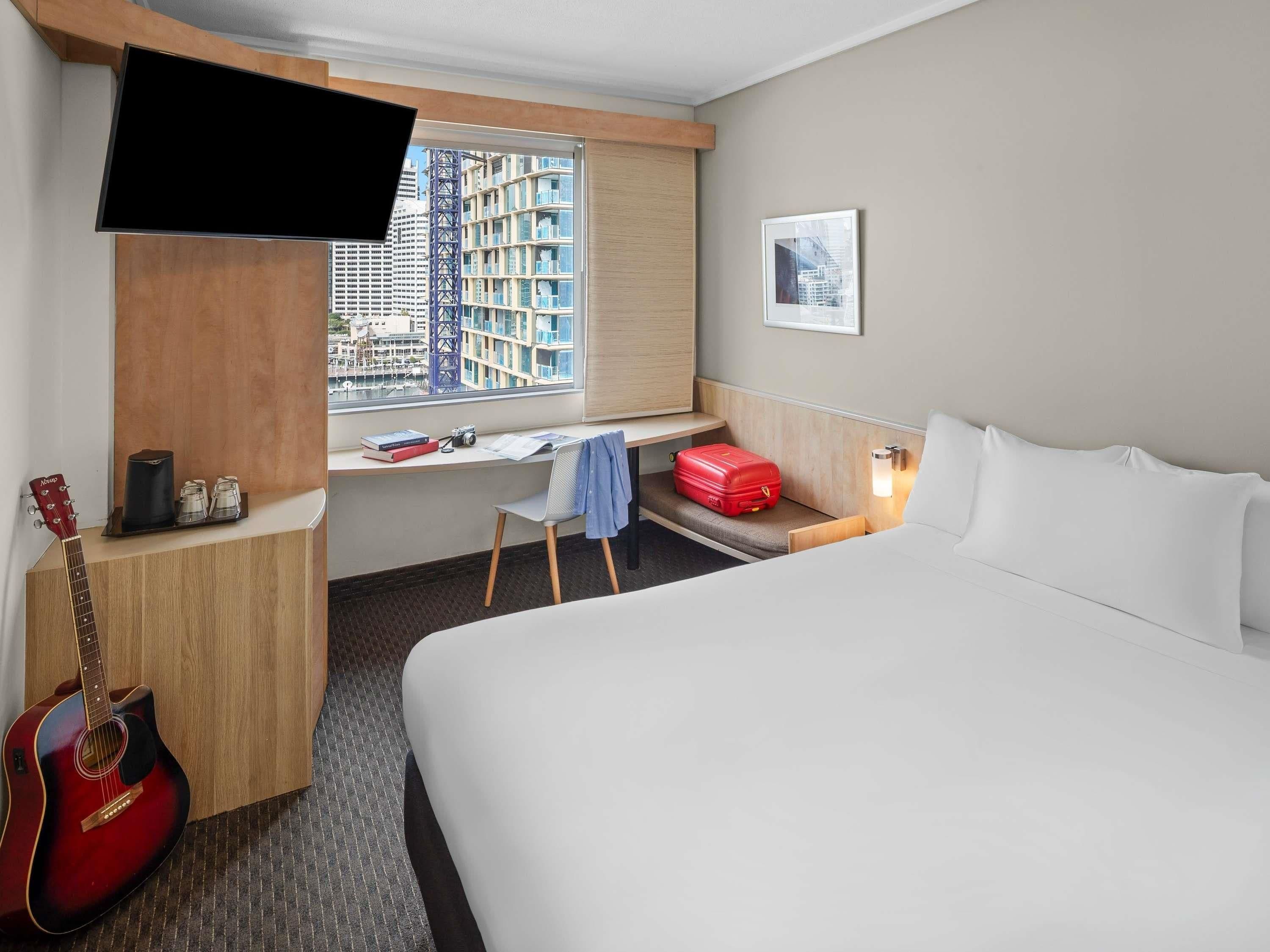 A hotel room with a bed, a desk with a chair, a guitar, a suitcase, and a TV mounted on the wall.
