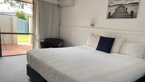 A neatly made bed with a white comforter and pillows, a chair, a painting on the wall, and a view through a window. - Macquarie Barracks Motor Inn (Port Macquarie)