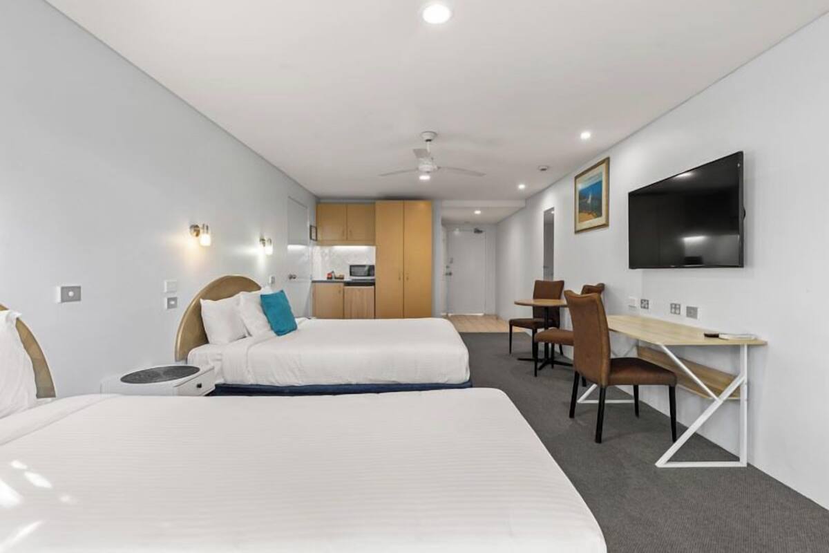 Quarto de hotel com cama, escrivaninha, cadeira, televisão e uma kitchenette ao fundo.