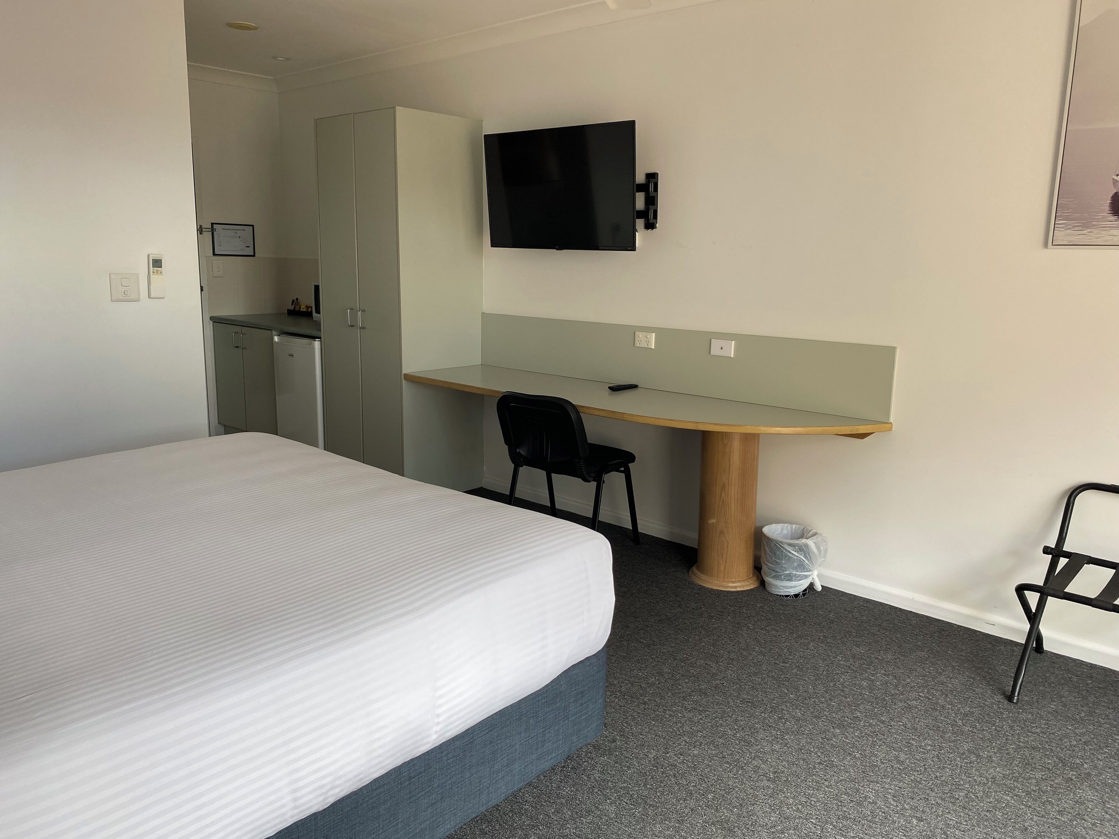 A hotel room with a bed, a desk with a chair, a television mounted on the wall, and a wardrobe.