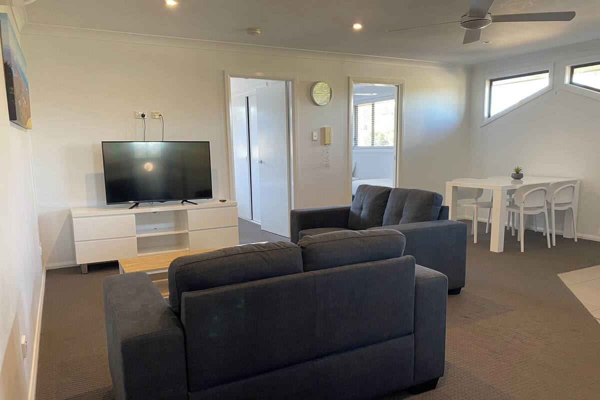 Uma sala de estar moderna com televisão de tela plana, sofás cinzas, uma mesa de jantar com cadeiras e um ventilador de teto.