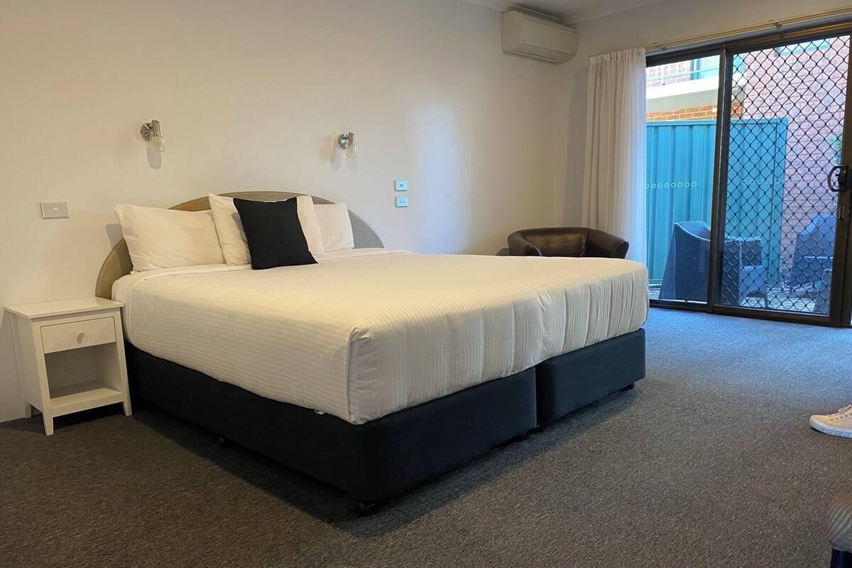 Quarto de hotel com uma cama grande, uma mesa de cabeceira, uma cadeira e vista por uma porta de vidro deslizante.