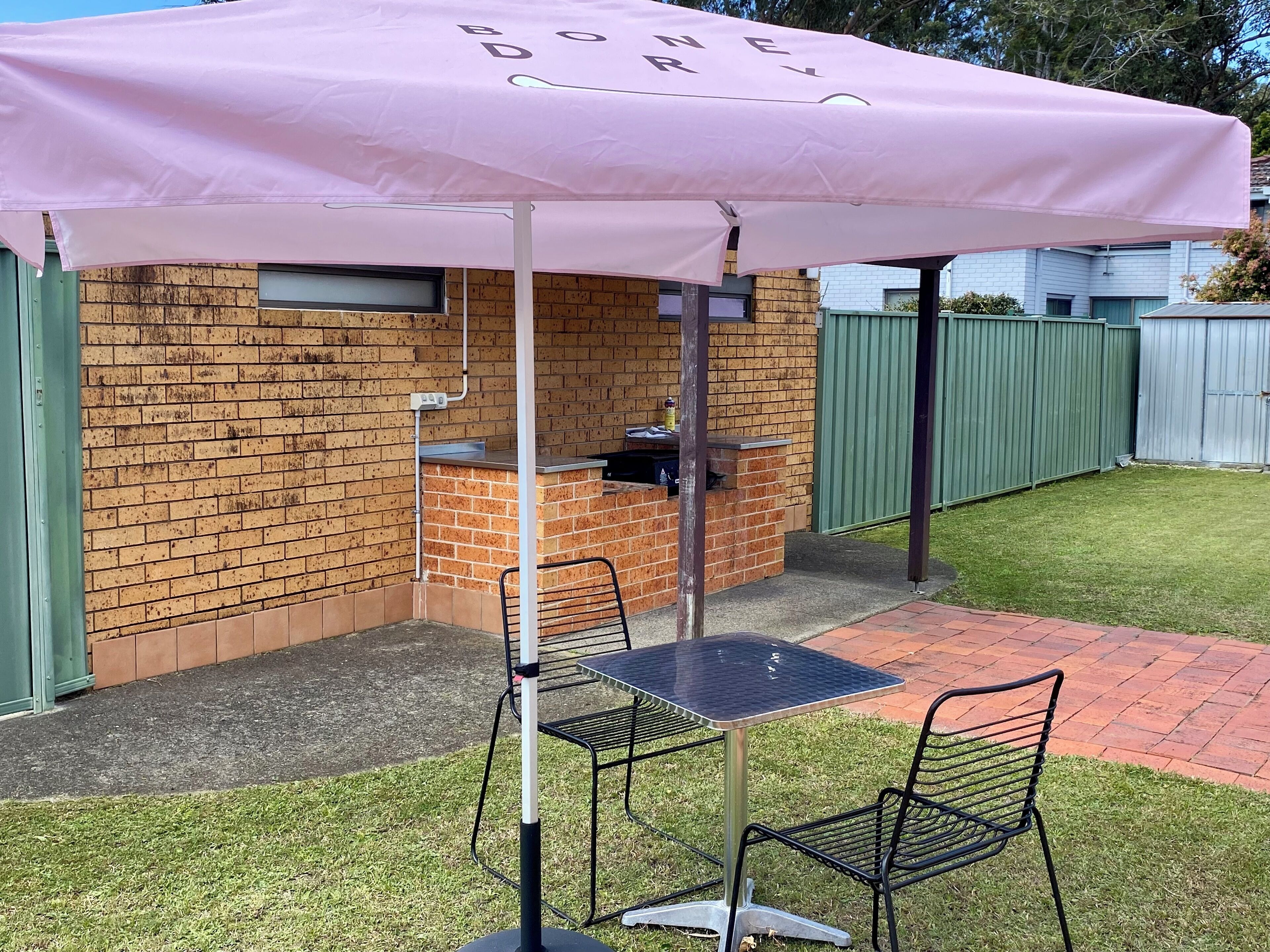 Zona de barbacoa de ladrillo con una mesa y sillas bajo un paraguas rosa.