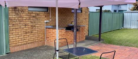 Zona de barbacoa de ladrillo con una mesa y sillas bajo un paraguas rosa.