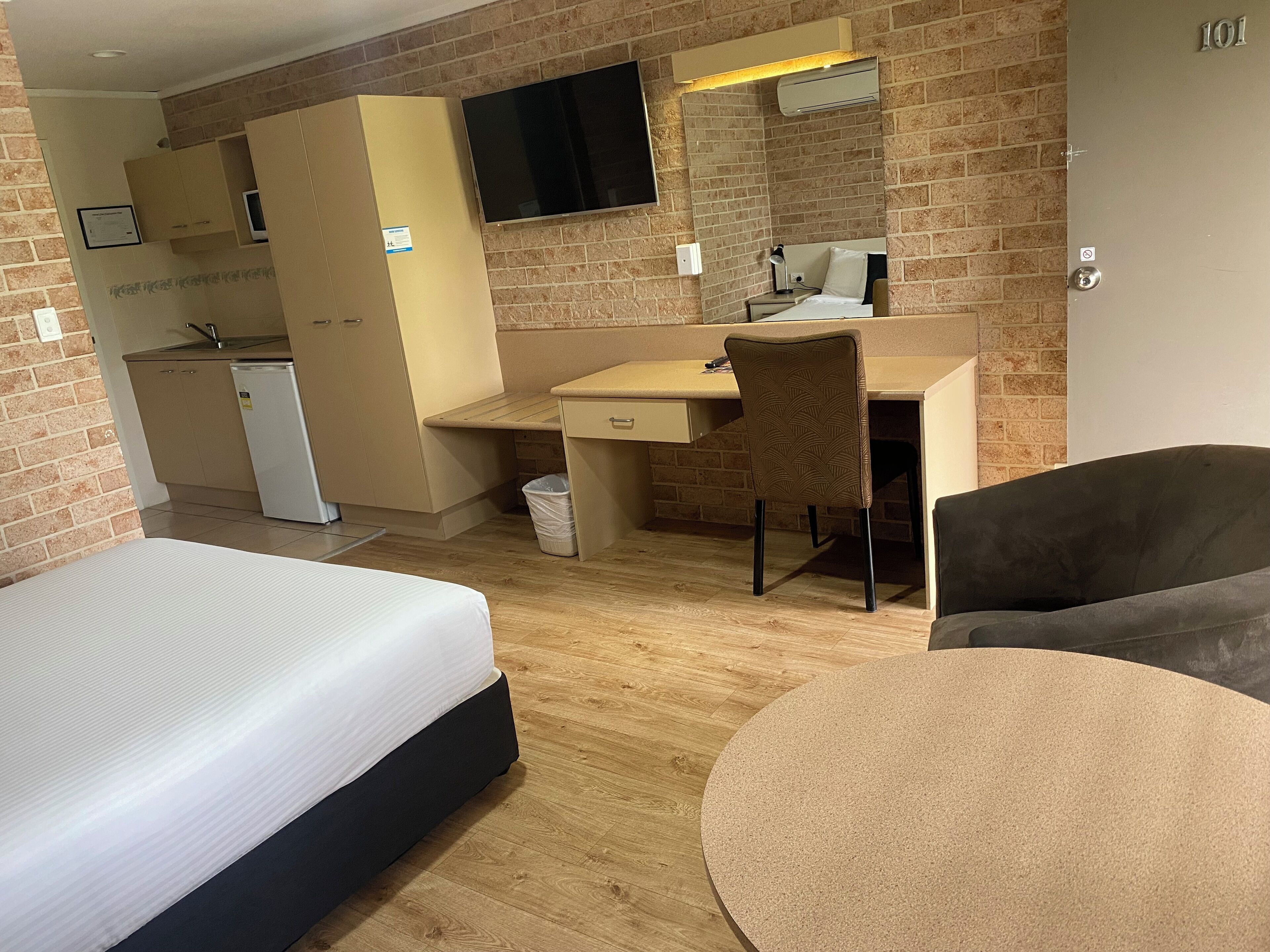 A hotel room with a bed, a desk, a chair, a round table, a TV, a small kitchenette, and a brick wall.
