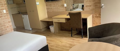 Una habitación de hotel con cama, escritorio, silla, mesa redonda, televisión, kitchenette pequeña y pared de ladrillo.