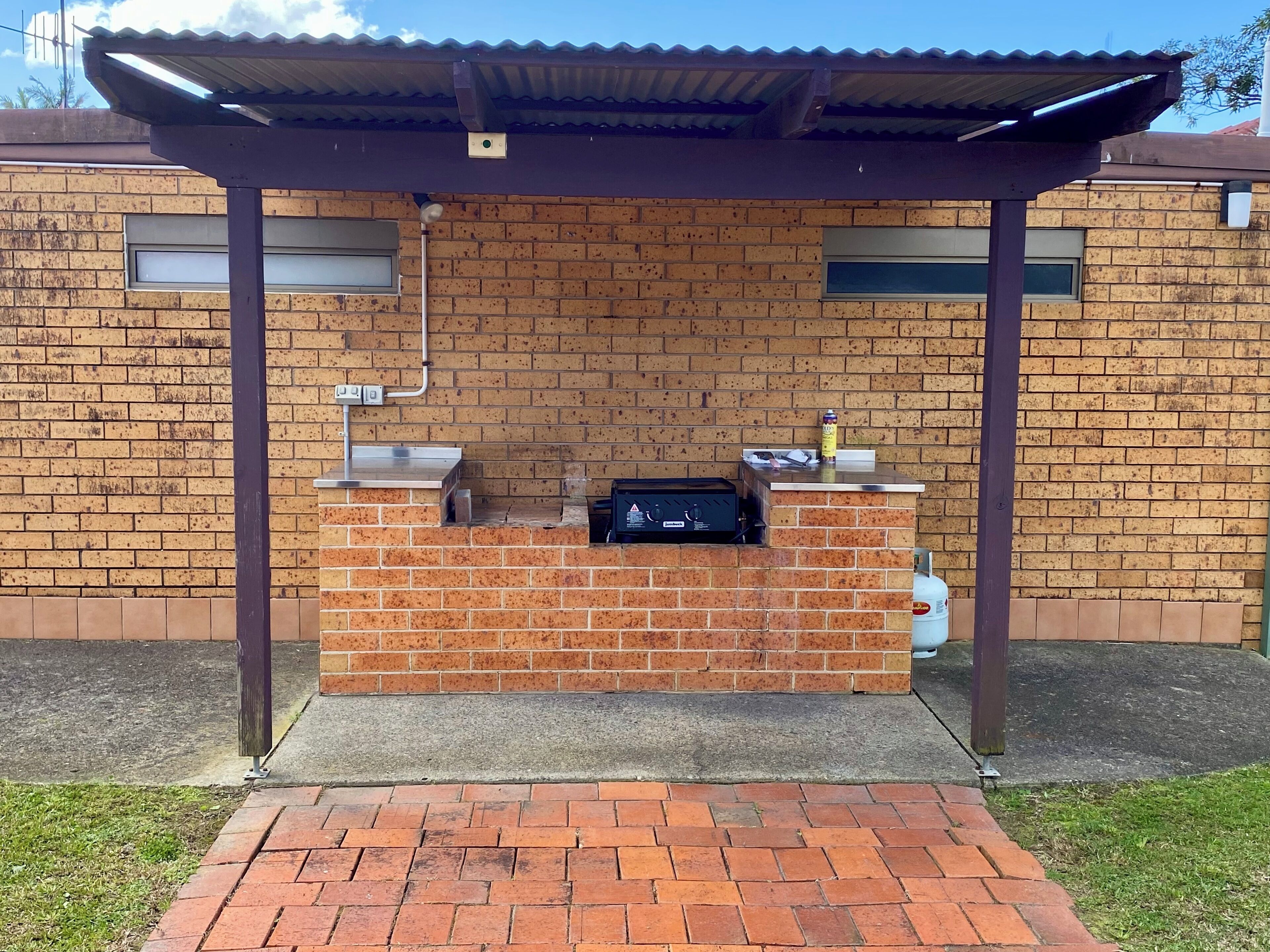 Un área de parrillada de ladrillo con una parrilla de metal, un tanque de gas y una lata de cerveza sobre el mostrador.