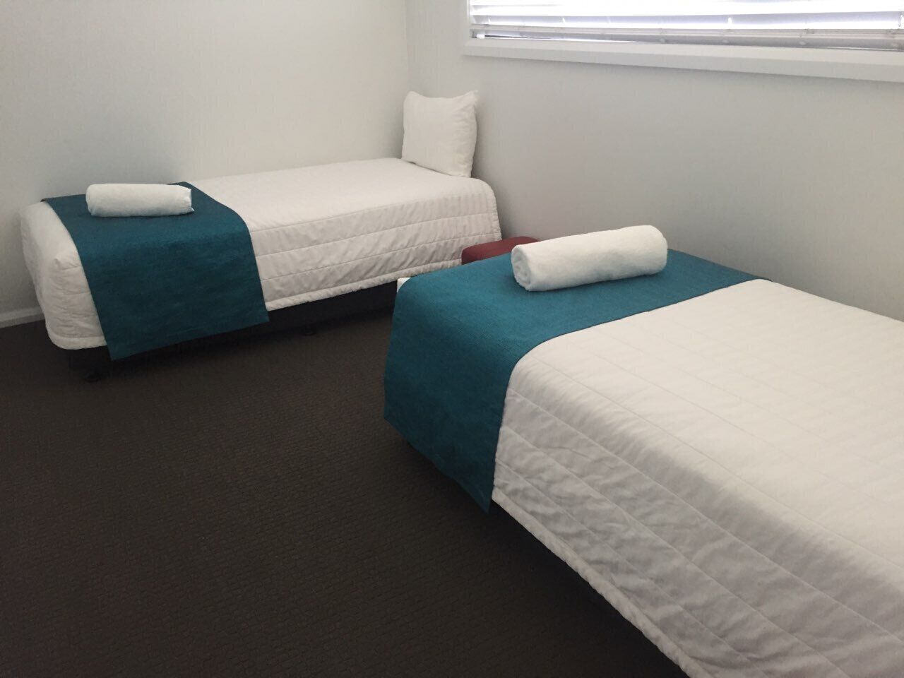 Two single beds with teal and white bedding, a red chair, and a window with blinds.