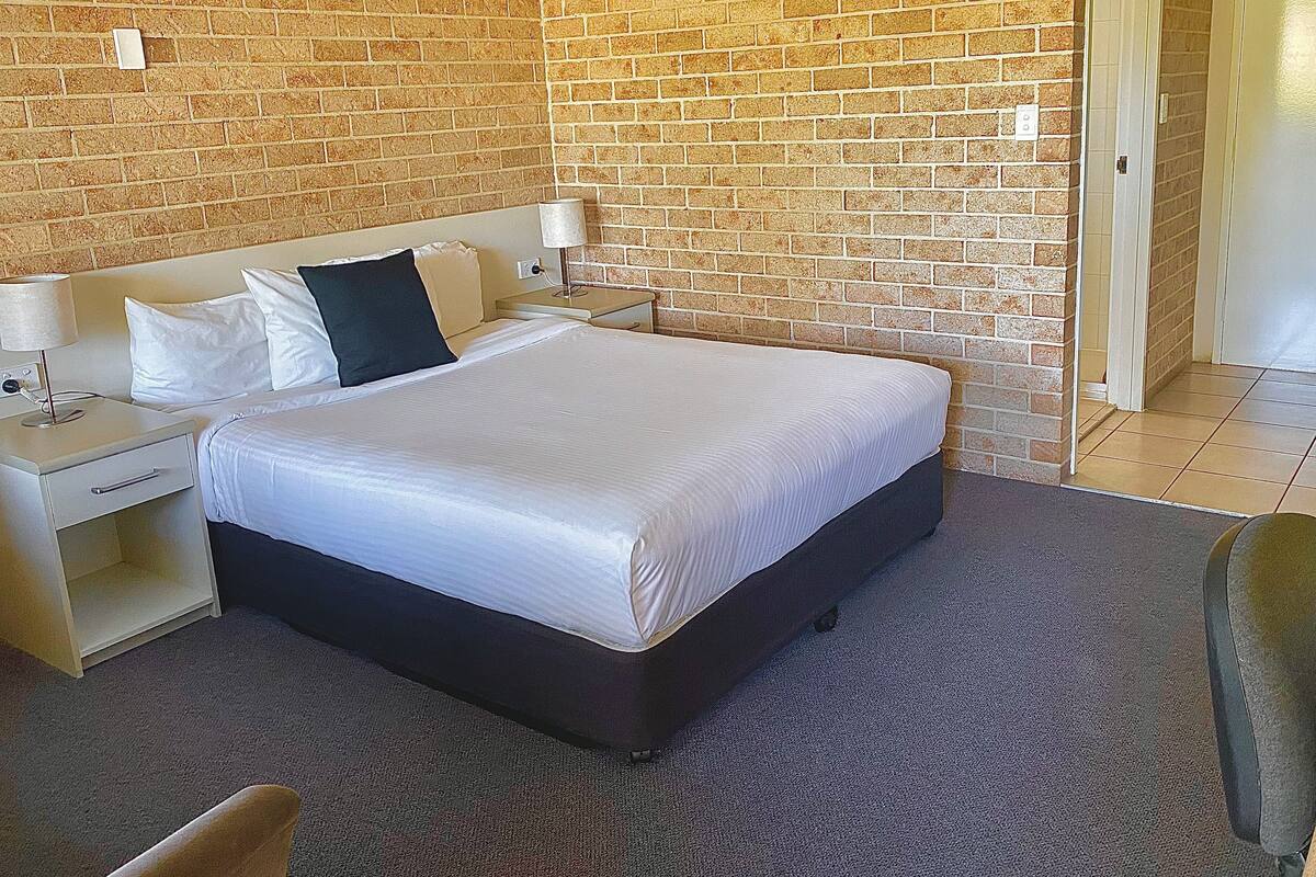 Quarto de hotel com parede de tijolos, cama com roupa branca, mesa de cabeceira, abajur e porta.