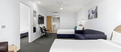 A hotel room with a large bed, a sofa, a desk, and a chair. There is a television on the wall and a ceiling fan.