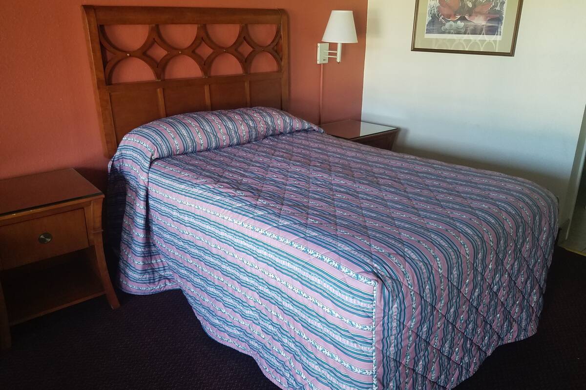 Un lit bien fait, recouvert d’une couvre-lit à motifs, avec une tête de lit en bois, une table de chevet dotée d’une lampe et un tableau encadré accroché au mur.