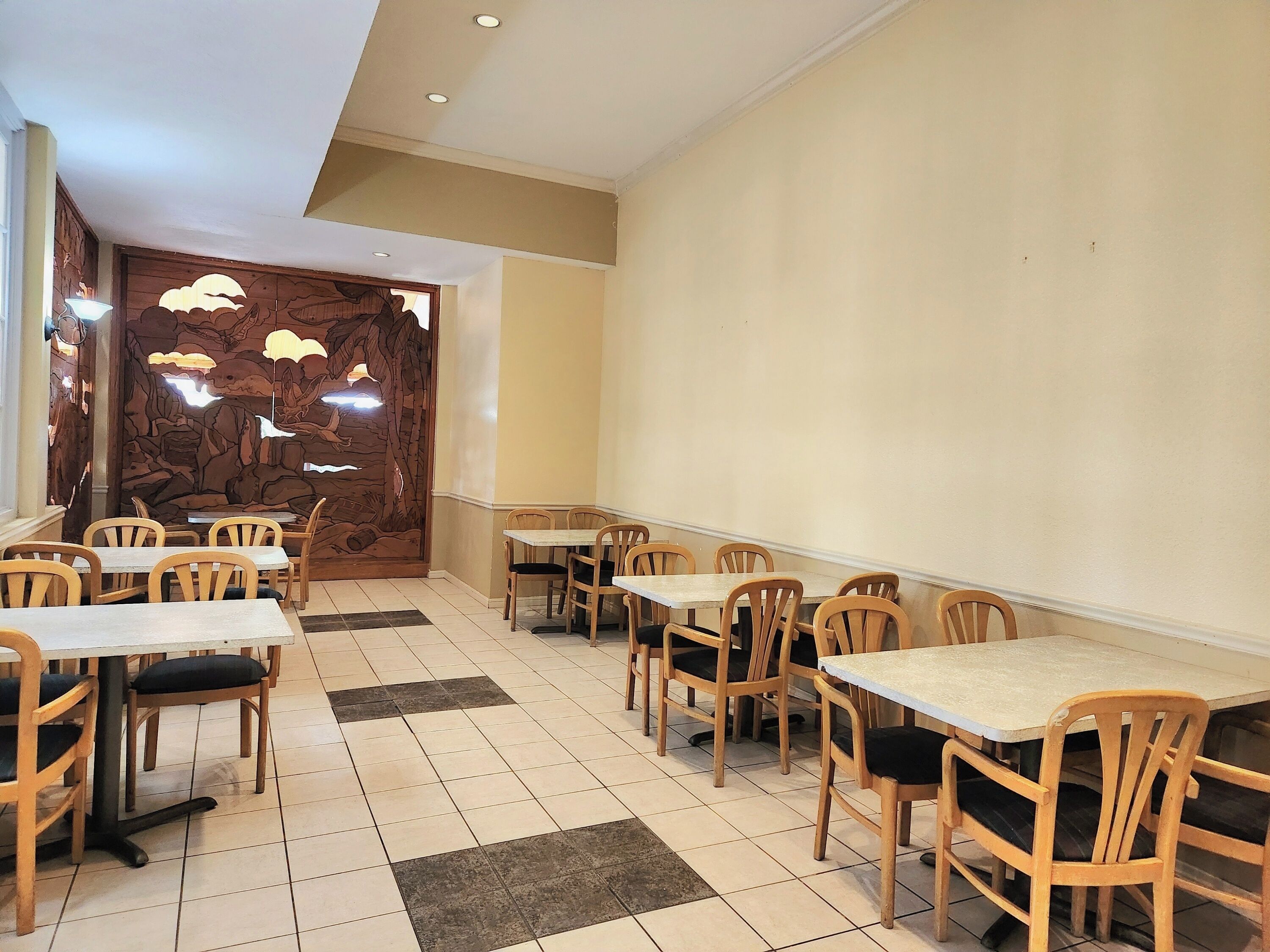 Un restaurante con mesas y sillas de madera, suelo de baldosas y una pared decorativa con un árbol y pájaros.