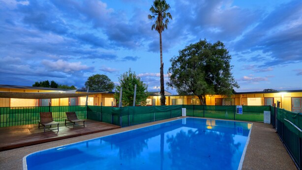 A swimming pool with a wooden deck and lounge chairs, surrounded by a green fence and trees. - Cootamundra Gardens Motel (Cootamundra)