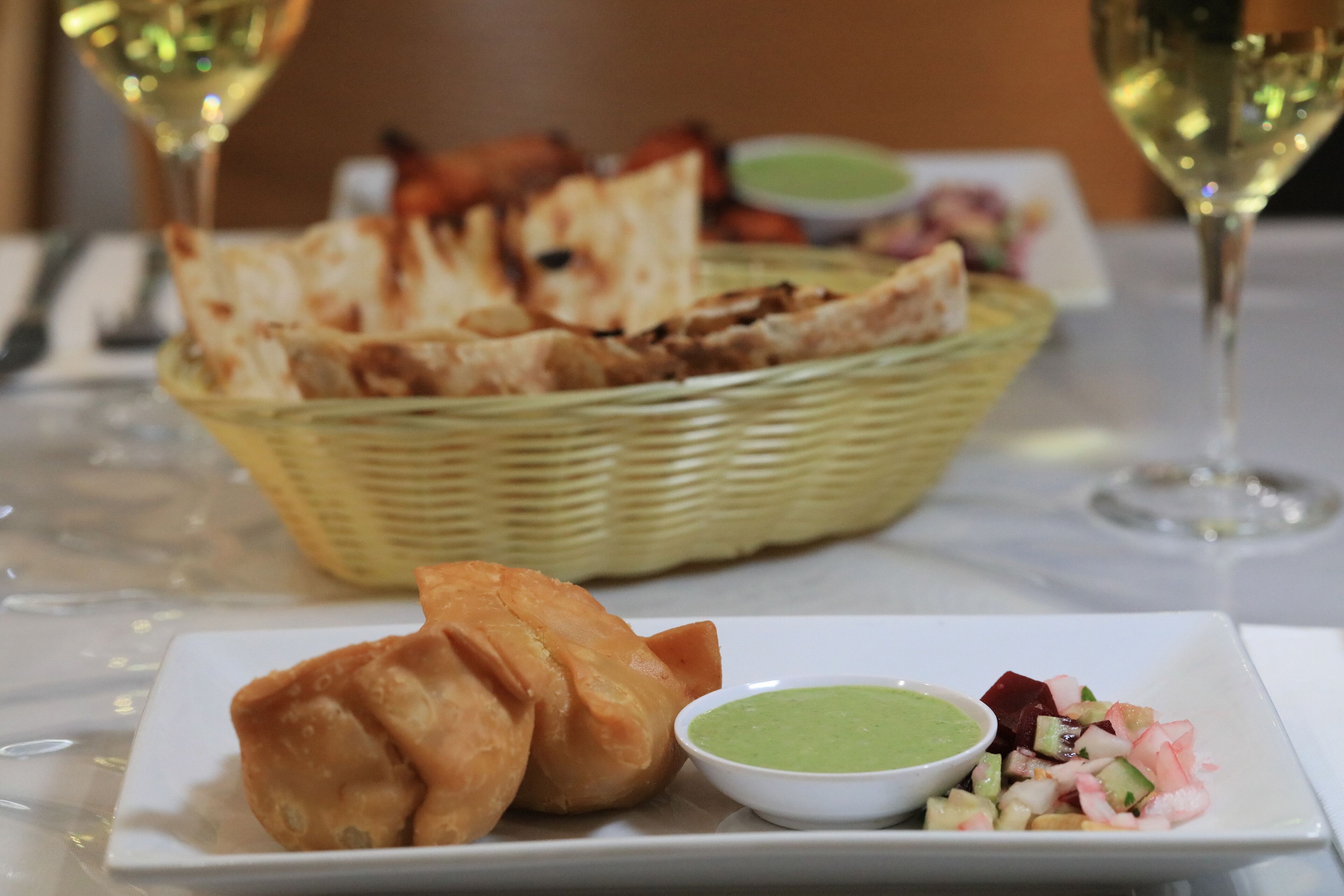 Un plato con samosas, salsa verde y ensalada de frutas, sobre una mesa con una cesta de pan y dos vasos de vino blanco.