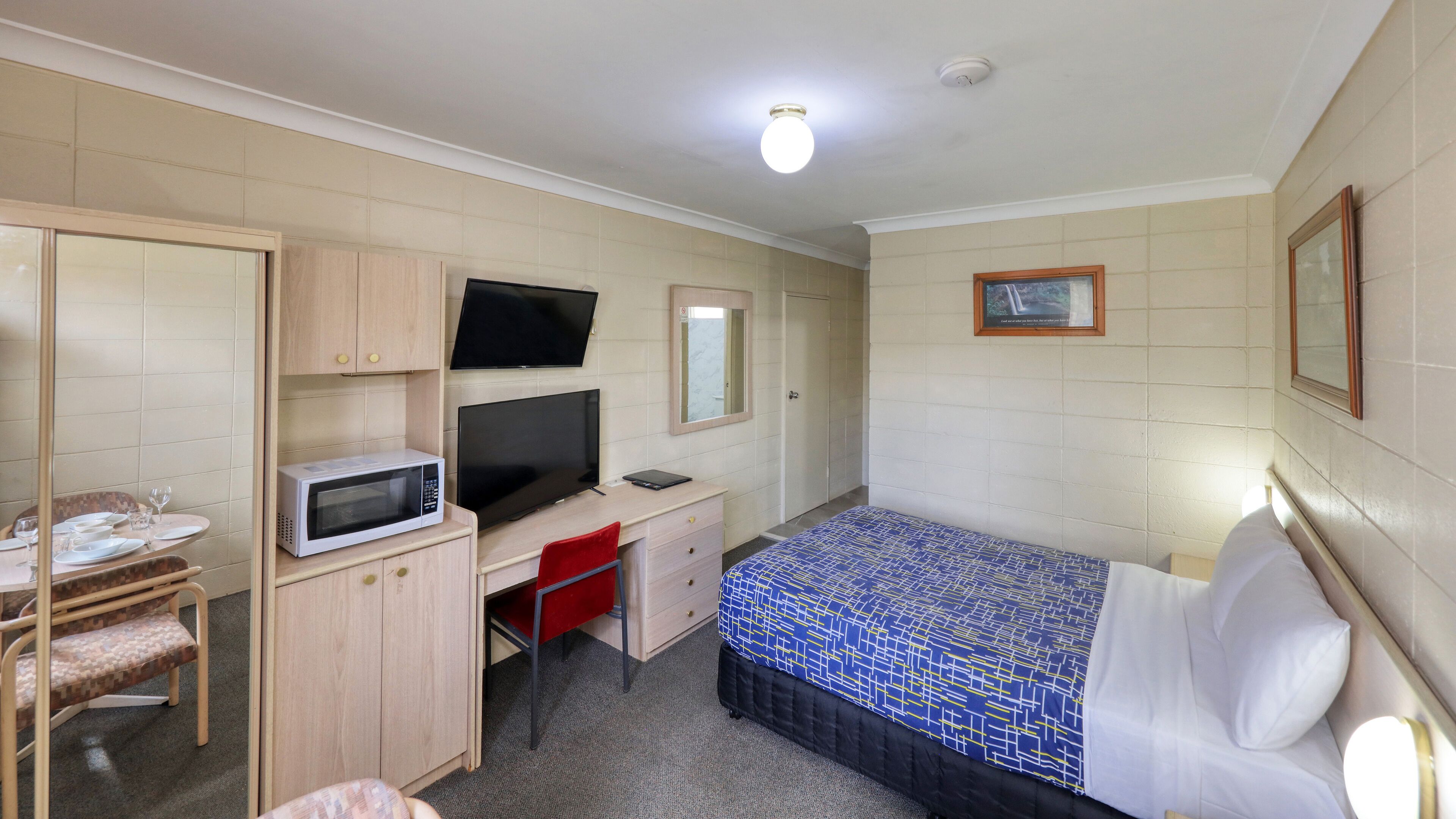 A hotel room with a bed, a desk with a computer, a microwave, a TV, and a small dining area with a chair and table.