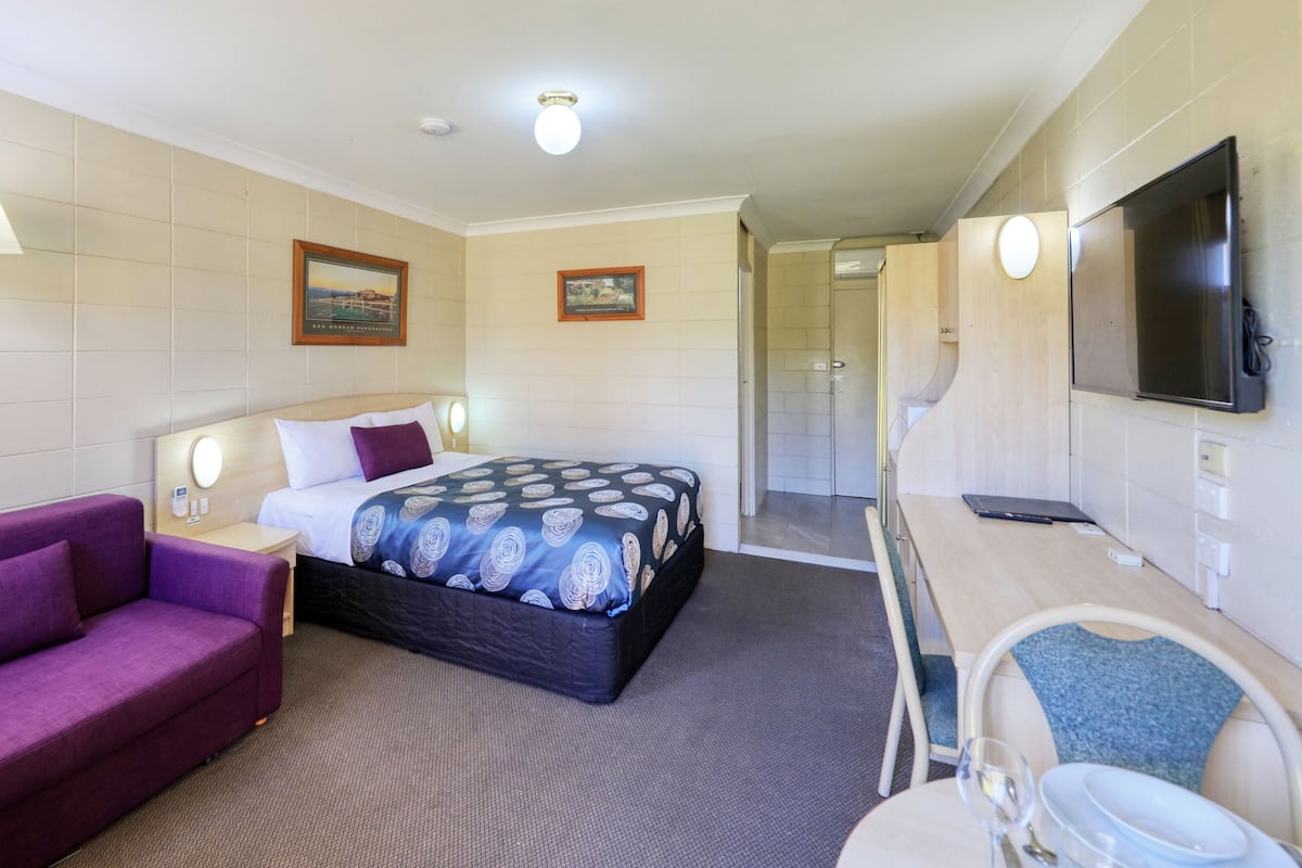 Une chambre d’hôtel avec un lit, un canapé violet, un bureau avec une chaise, une télévision et une salle de bain visible à travers une porte ouverte.