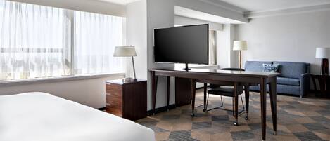 A hotel room with a bed, a desk with a television, a sofa, and a window with sheer curtains.