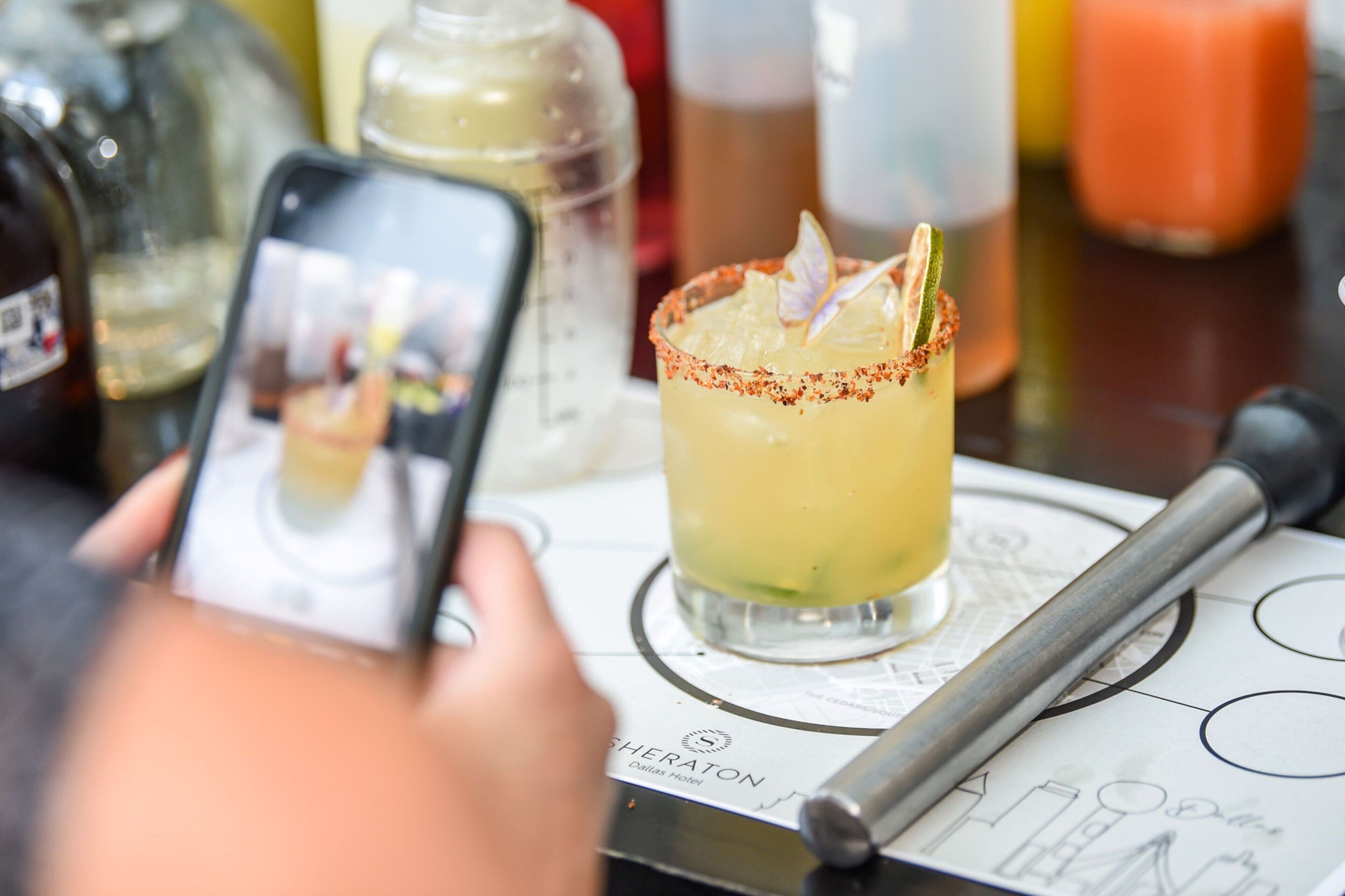 A cocktail glass with a garnish on a bar counter, a smartphone displaying a photo of the drink, and a menu with the name "Sheraton Dallas Hotel".
