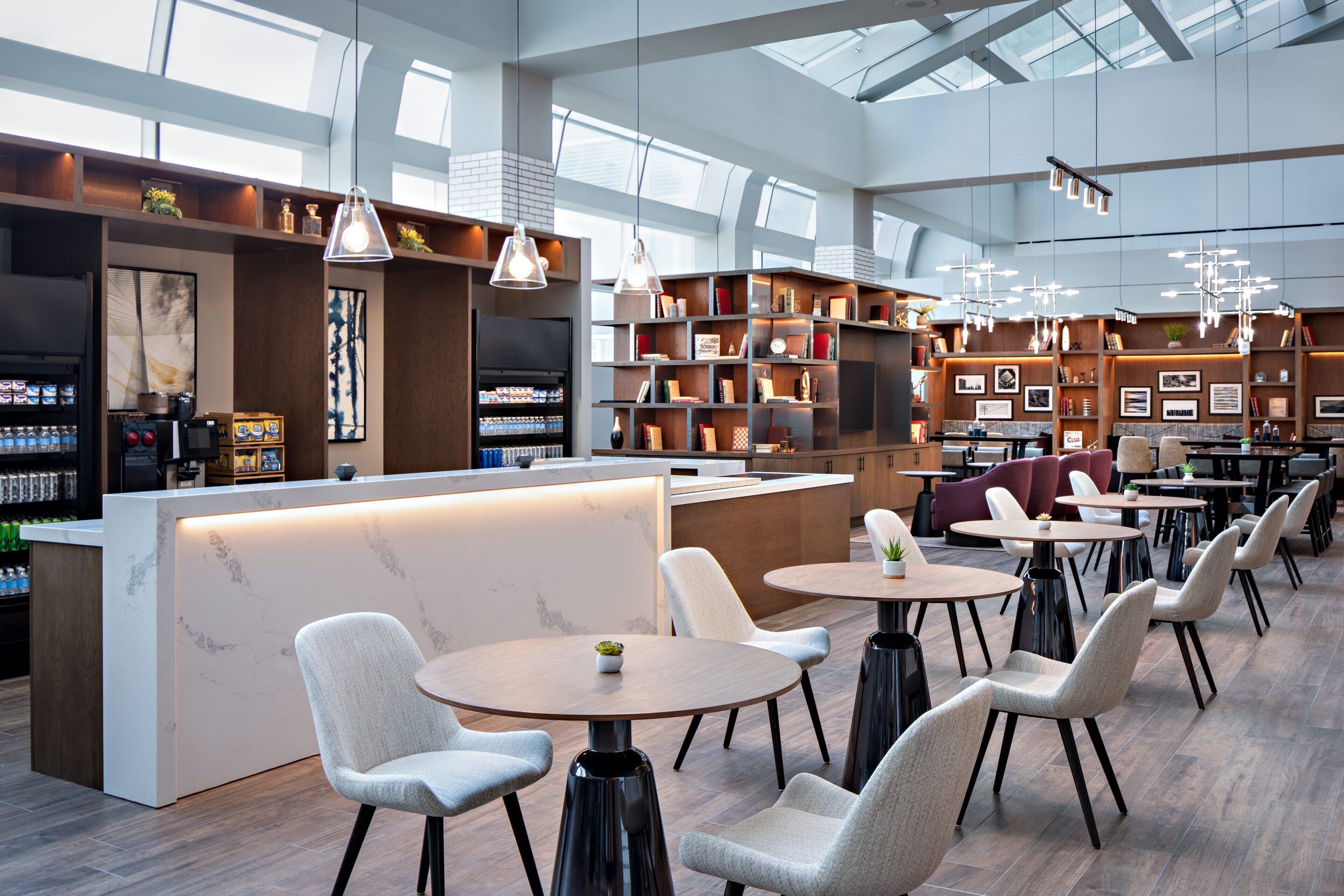 A modern cafe with a central counter, multiple seating areas, and a large windowed wall.